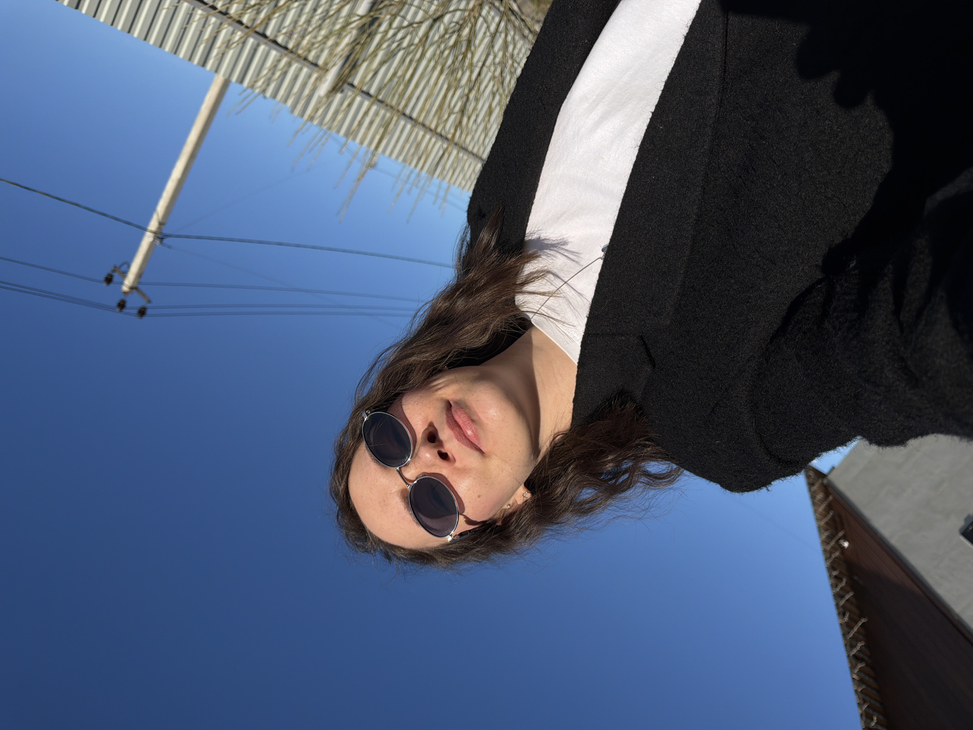 The image is an angled low-angle shot of a woman wearing round sunglasses, a white shirt, and a black blazer. Her hair is wavy and brown. The background is primarily a clear blue sky. In the upper left corner, there is a utility pole with power lines extending across the frame. There is also a glimpse of a corrugated metal roof. In the lower right corner, part of a building is visible. Based on location information, the picture may be set in Chaikovsky, Russia.