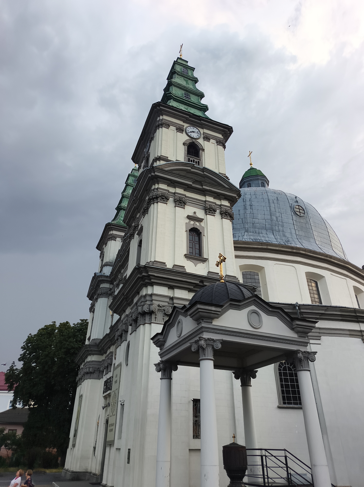A low-angle, full shot captures the exterior of the Ternopil Cathedral in Ternopil, Ukraine, under a cloudy sky. The cathedral features a white facade with grey detailing, green tiered spires, and a large, blue-grey domed roof. A golden cross adorns the top of the tallest spire and a smaller dome. A portico with white columns and ornate capitals stands in the foreground, leading to a staircase. Two people are visible in the lower left of the frame, looking up at the cathedral. Trees and a section of another building are visible to the left of the cathedral.