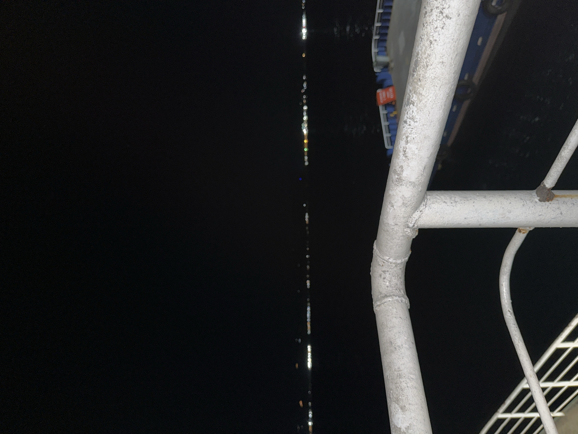 The image depicts a dark, nighttime scene, likely captured from the deck of a boat or ferry in Davao City, Philippines. The foreground is dominated by a sturdy white metal railing, showing signs of weathering with specks of dirt and what appears to be minor rust near a joint. To the right, another white metal bar forms a part of this railing structure, curving downwards.
Behind the railing, in the upper right, a blurred blue object is visible, possibly part of the vessel's structure or seating. A small, indistinct red rectangular element with unreadable white text is attached to this blue object.
The majority of the background is pitch black, suggesting open water or sky at night. A striking feature is a vertical line of small, blurred, bright lights that extends from the top to the bottom of the left side of the frame. These lights, appearing as bokeh, vary in color, showing primarily white, but also hints of yellow, orange, and blue. They are likely reflections of distant lights or the vessel's own lights on the water's surface, indicating a calm body of water. The overall scene suggests a quiet journey by water during the night. No specific activity beyond being onboard a vessel is discernible.