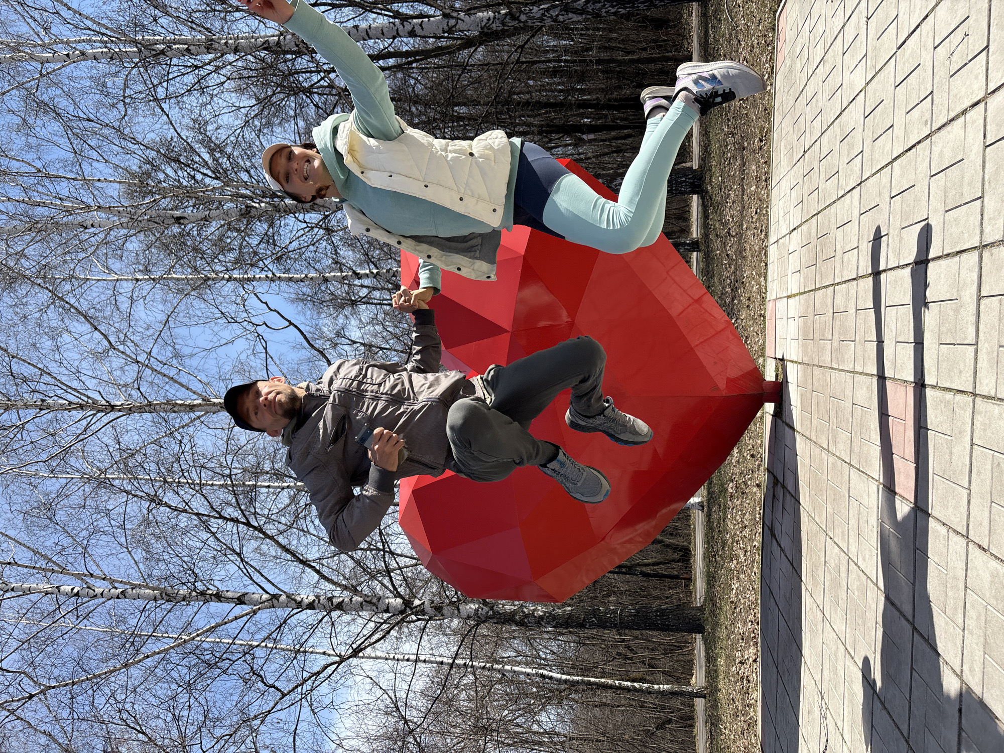 This outdoor image, taken in Ulyanovsk, Russia, captures two individuals, a man and a woman, playfully posing next to a large, red, faceted heart-shaped sculpture. The scene suggests a bright, sunny day in late winter or early spring, characterized by a clear blue sky and bare birch trees in the background, their white trunks standing out against the sky.
The woman, positioned higher, appears to be "floating" or jumping, holding hands with the man below. She is dressed in a light blue long-sleeved top under a white puffer vest, light blue leggings with a dark blue upper section, and grey and white New Balance sneakers. She wears a light-colored cap and has a joyful expression.
The man, also seemingly defying gravity, is dressed in a grey jacket, dark trousers, grey sneakers, and a dark baseball cap. He is holding a dark-colored smartphone in his left hand. Both individuals cast long, distinct shadows on the light-colored paved ground beneath them, indicating the sun is overhead, likely midday. The large red heart sculpture dominates the lower-middle portion of the image, providing a vibrant focal point. The overall mood is lighthearted and energetic, showcasing a moment of playful interaction in a park-like setting. No other significant text is visible.