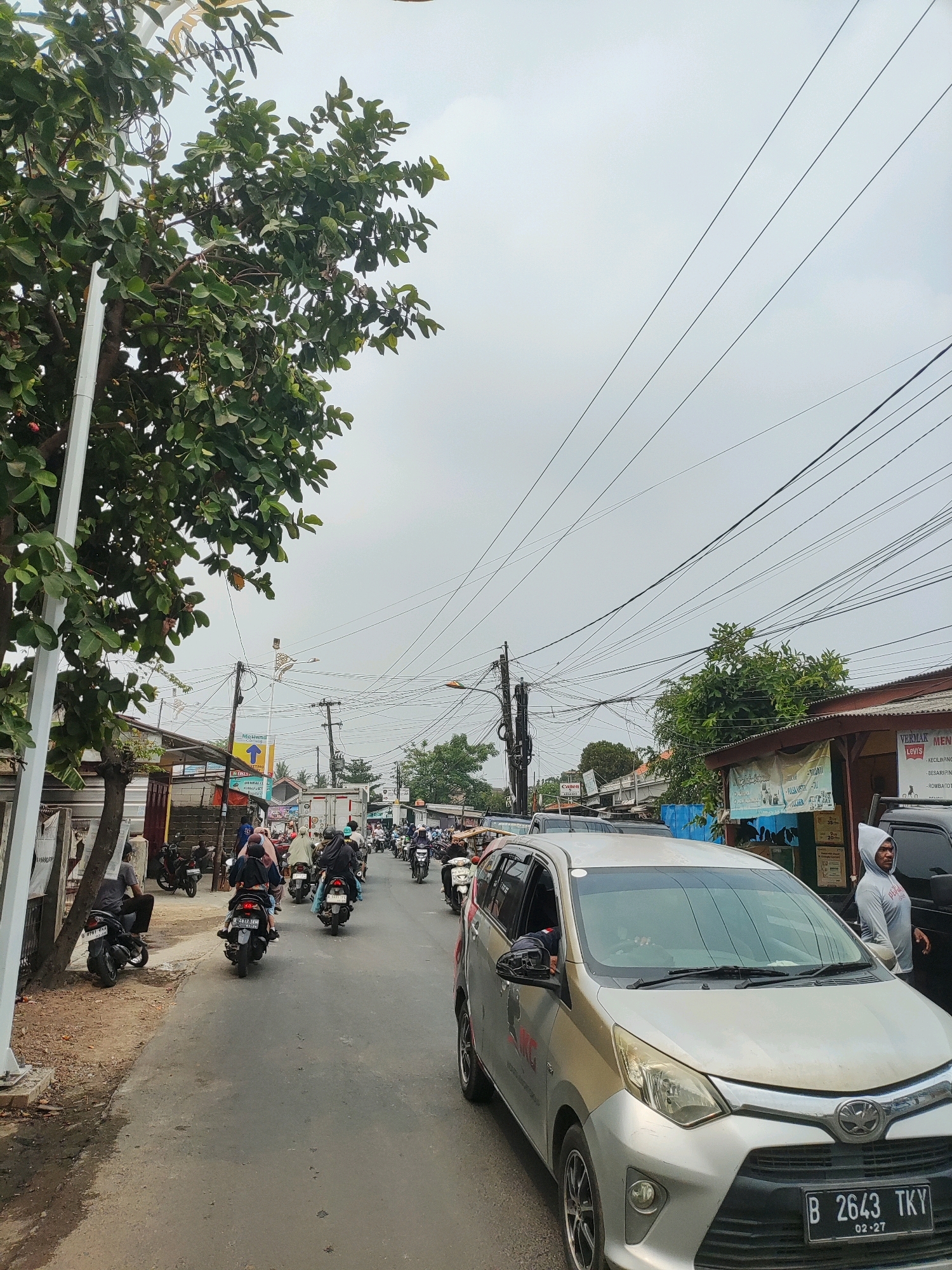 The image shows a street scene in Wanajaya, Indonesia, on a cloudy day. The foreground features a silver car with a license plate reading "B 2643 TKY 02-27". A person wearing a grey hooded sweatshirt stands next to the car. To the left of the car is a tree with dense green foliage and visible branches.
The street is lined with buildings and shops on both sides, and there is a significant amount of overhead electrical wiring. Traffic is moderate, with several motorcycles and a few cars on the road. People on motorcycles are seen riding in both directions. Some buildings have signs, including one with the text "VERMAK MEN" and "LEVIS". The road surface is asphalt. The overall impression is of a busy urban or semi-urban environment with typical Indonesian street activity.
