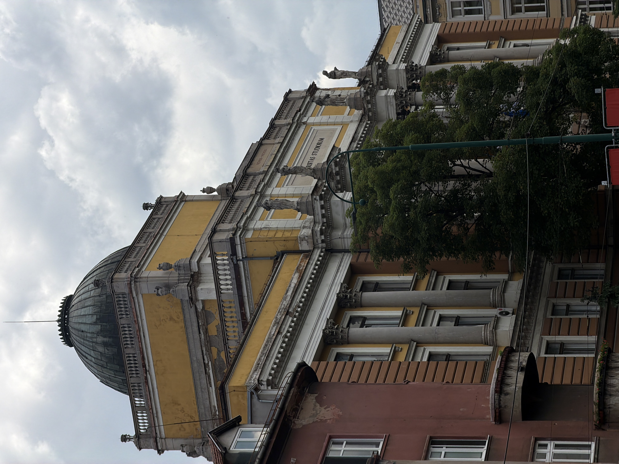 The image shows the National and University Library of Bosnia and Herzegovina in Sarajevo. The building, constructed in the Neo-Moorish style, features a prominent dome, arched windows, and ornate architectural details. The façade is adorned with statues and a large inscription that reads "UNIVERSITAS STUDIORUM". The surrounding area is characterized by other buildings and lush green trees, with a cloudy sky overhead. The library's yellow and white exterior contrasts with the reddish-brown building to its left. A green lamppost stands in the foreground.