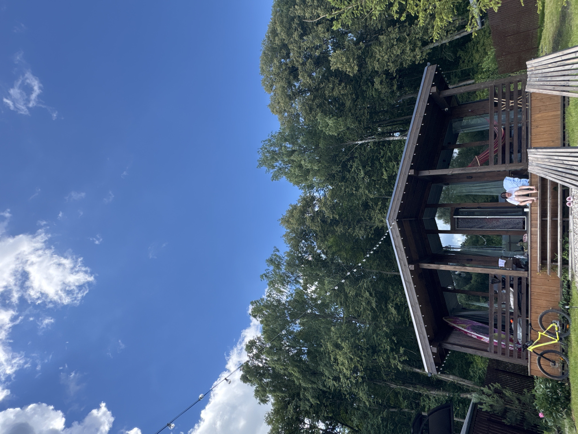 A bright, sunny day with scattered clouds is captured in this vertical outdoor shot. In the foreground, a wooden house with a sloping roof and large glass windows sits nestled among lush green trees. A woman in a white t-shirt and blue shorts is seated on the wooden steps leading up to the house, her legs bent and feet bare. A bright yellow bicycle is parked on the grass to the left of the house, leaning against a wooden railing. A surfboard is propped against the side of the house, and a red hammock is visible on the porch. The scene appears to be a tranquil retreat, possibly in the city of Serpukhov, Russia, given the context provided. The overall impression is one of leisure and relaxation in a natural setting.