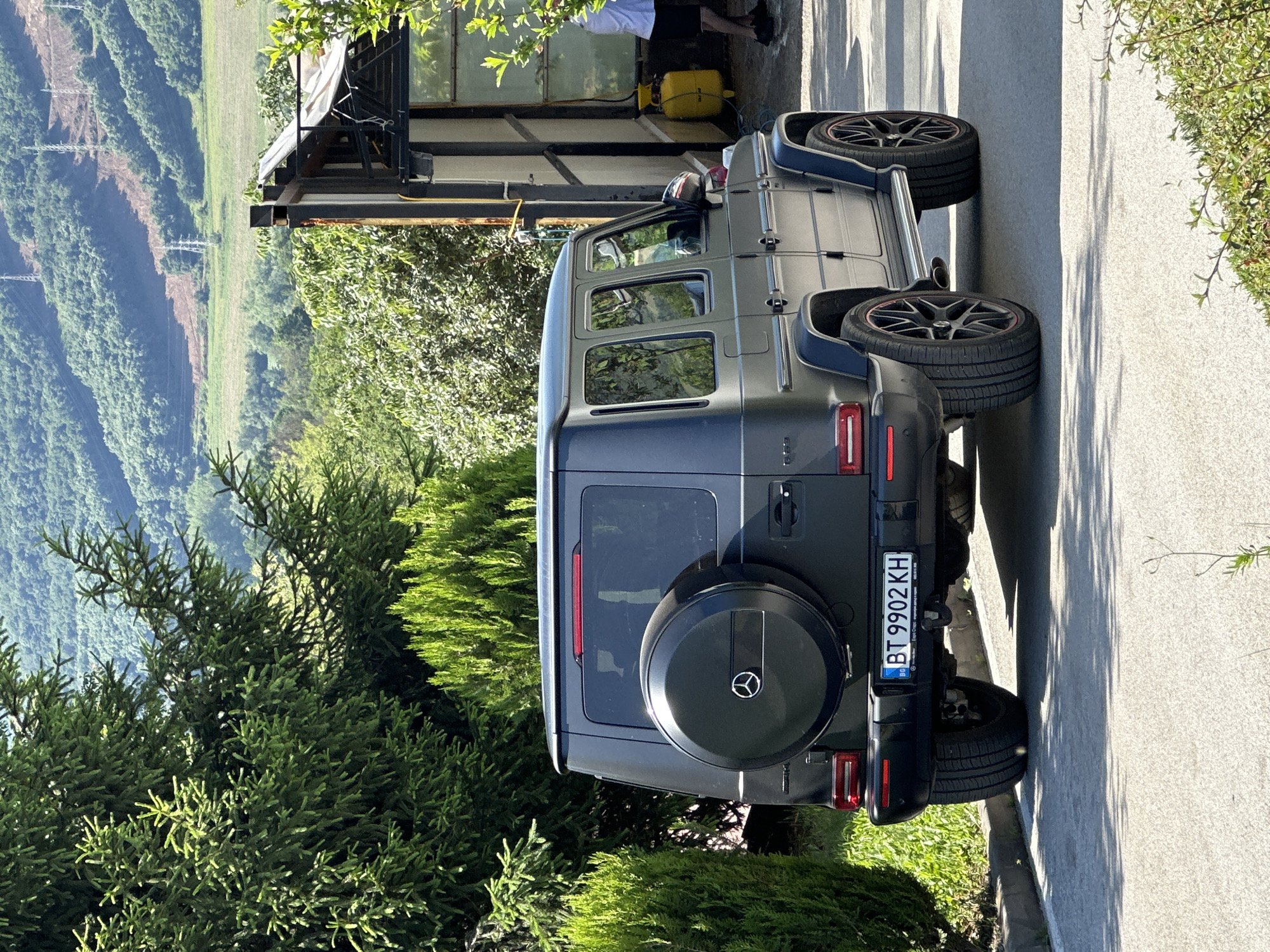 A gray Mercedes-Benz G-Class SUV is parked on a paved surface in Ledenik, Bulgaria. The vehicle has a spare tire mounted on the rear door and a Bulgarian license plate reading "BT 9902 KH". A person in a white shirt is visible near the SUV. Behind the vehicle, a structure with a metal frame stands adjacent to lush greenery. A hillside and mountains are visible in the distance. The scene is well-lit with natural sunlight.