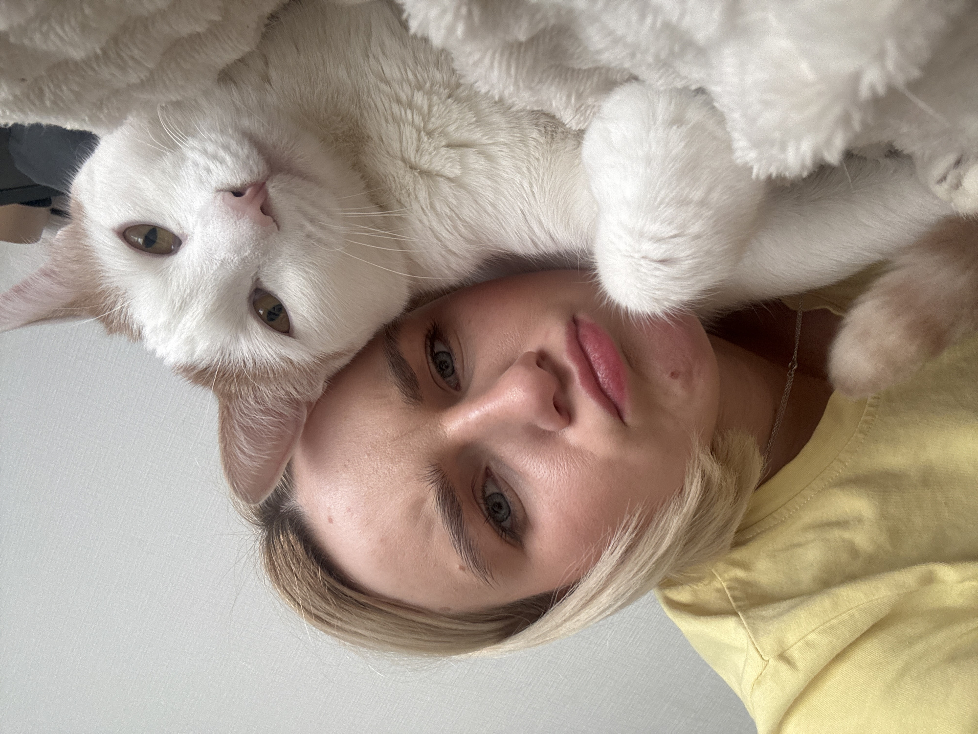 A woman with blonde hair wearing a yellow t-shirt is photographed with a white and orange cat. The cat is lying on top of the woman's head and shoulders. The photograph appears to be taken from an upside-down perspective. No information suggests this photo was taken in Vsevolozhsk, Russia.