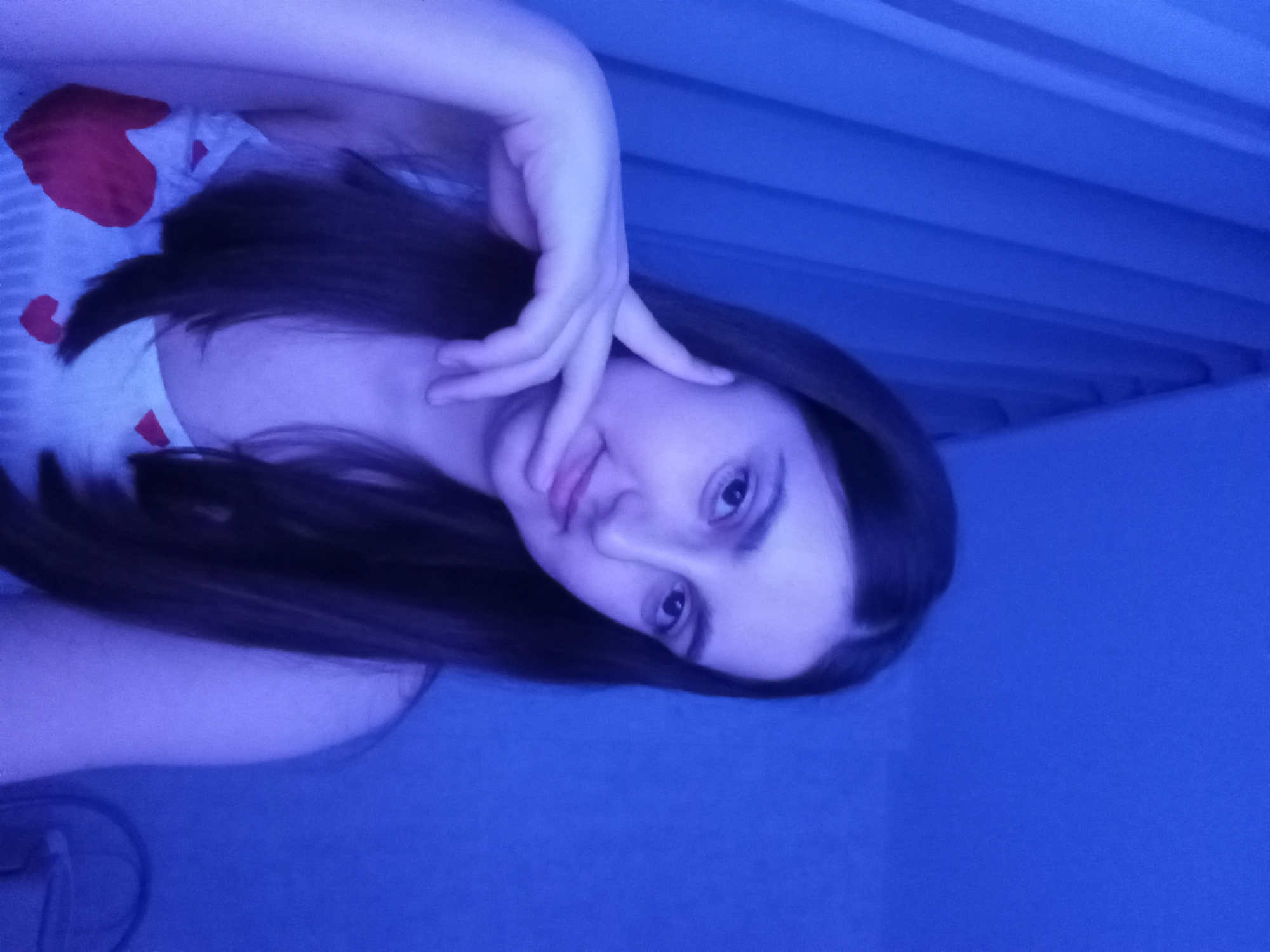 A young woman with long, dark hair is shown in a close-up, selfie-style shot. She is wearing a white top with red hearts. Her left hand is resting against her face, with her fingers gently touching her cheek and chin. The background consists of blue curtains, suggesting an indoor setting. The lighting is predominantly blue, casting a cool tone over the entire image. The woman's expression is neutral, with her gaze directed towards the camera. There are no identifiable landmarks or contextual clues in the image that would suggest a specific location within Krasnodar, Russia.
