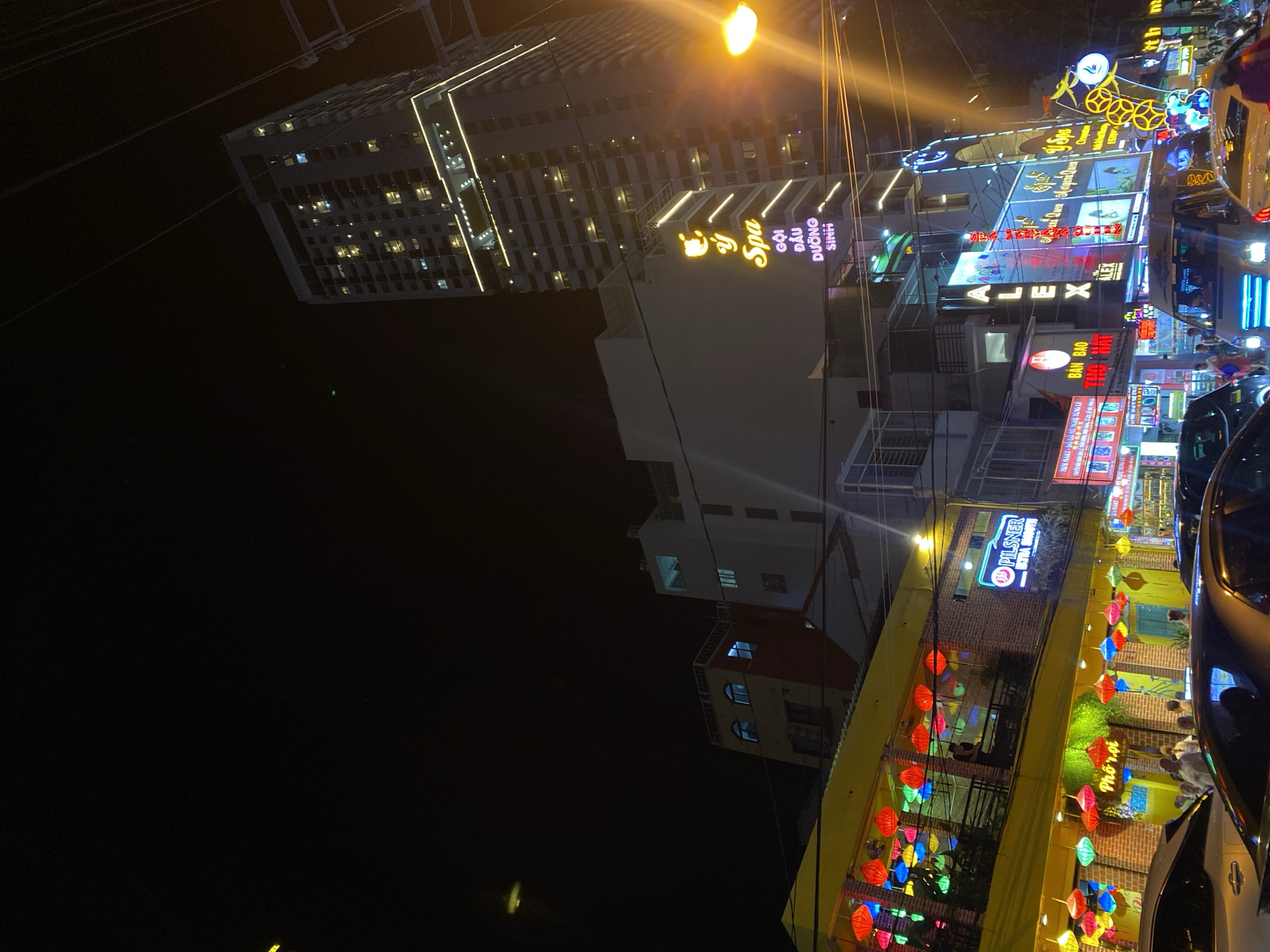 The image captures a vibrant street scene at night in Nha Trang Ward, Vietnam. The primary subjects are the illuminated buildings and signage that line the street, with several cars parked and driving along it. The scene is filled with neon lights and colorful decorations, including many paper lanterns strung across the front of a yellow building, suggesting a festive atmosphere.
One prominent building features a large sign for "Ý Spa" with the words "GỘI ĐẦU DƯỠNG SINH" below it. Further down the street, other signs are visible, including "ALEX" in large letters and a "PILSNER" sign with "EXTRA SMOOTH" underneath. A "MINI MART - ĐỒ GỖ MỸ NGHỆ" sign is also present. The yellow building adorned with lanterns appears to be a restaurant or cafe, with the Vietnamese phrase "Phố rọi" visible.
The time of day is clearly night, indicated by the darkness and the prominence of artificial lighting. There are some people visible, blurred by the low light and motion, suggesting a busy evening. The weather appears to be clear, as there are no indications of rain or adverse conditions. The overall impression is one of a lively urban environment at night, bustling with activity and illuminated by a variety of colorful signs and decorative lights.