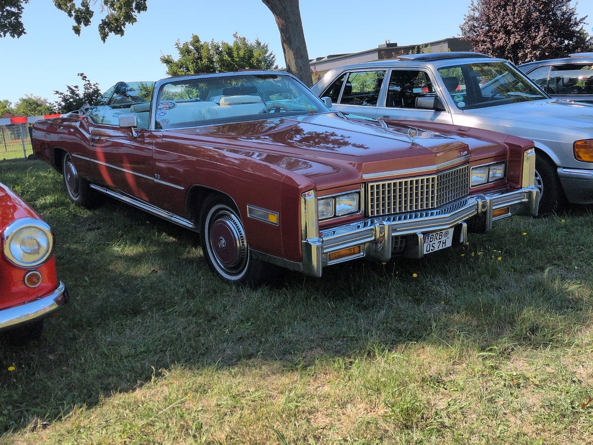 Two classic convertible cars are parked side-by-side on a grassy area in Hohenferchesar, Germany. The car in the foreground is a dark red Cadillac Eldorado convertible, with its top down, revealing a white interior and a tan steering wheel. It has a chrome grille, rectangular headlights, and a license plate that reads "BRB OS 7H". To the right of the Cadillac, a light silver sedan, likely a Mercedes-Benz, is parked. Its windows are tinted, and a portion of its hood and roof are visible. A red car is partially visible on the far left of the image. The cars are situated on a lawn with scattered yellow flowers and under the shade of trees. The sky is clear and blue.
