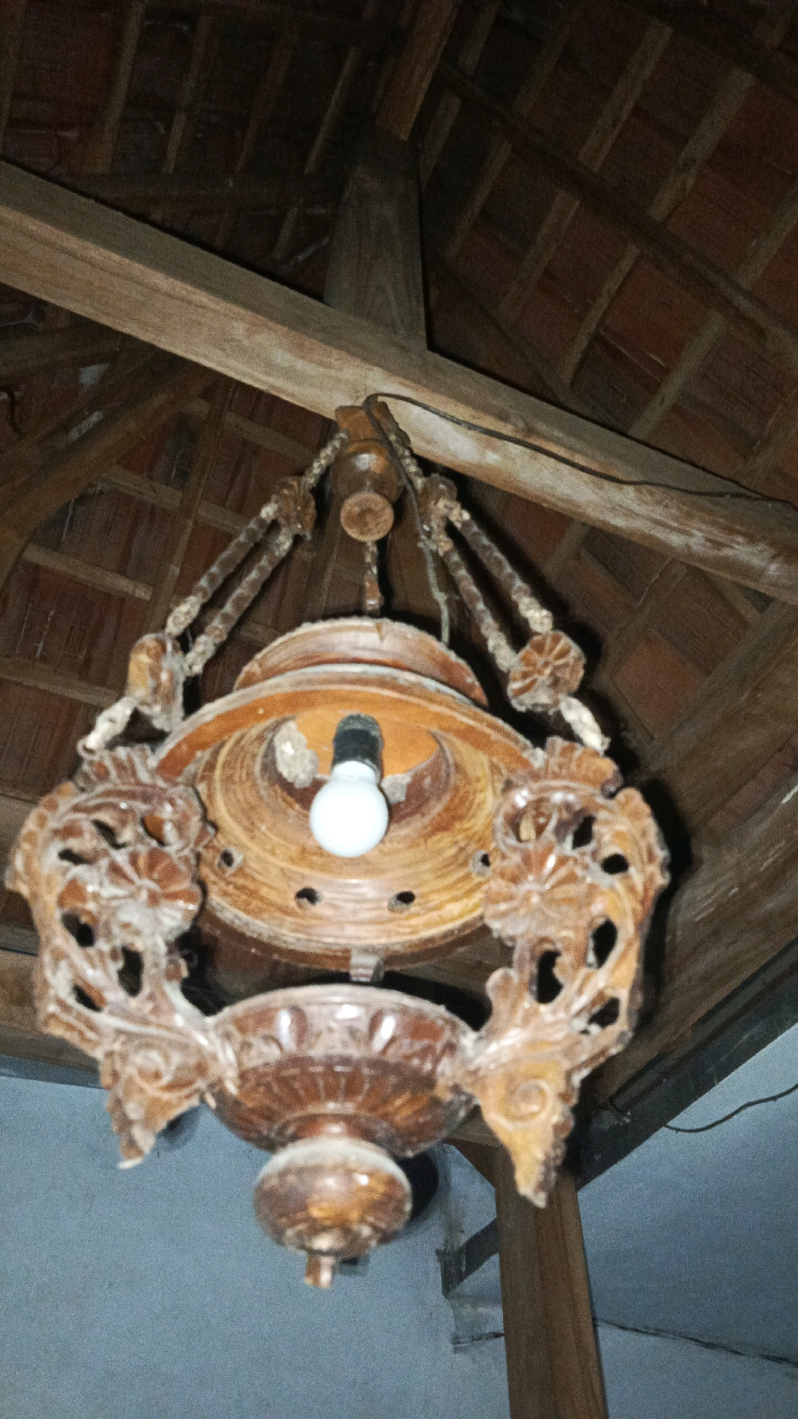 The image shows a traditional wooden chandelier hanging from a wooden ceiling with tiled roofing. The chandelier is intricately carved and features a light bulb. The environment appears to be indoors, possibly a home or a traditional building, with visible wooden beams and roof tiles. The overall scene evokes a rustic and perhaps aged ambiance. The specific location in Babadan, Indonesia, suggests a cultural context for this type of ornamentation and architecture.