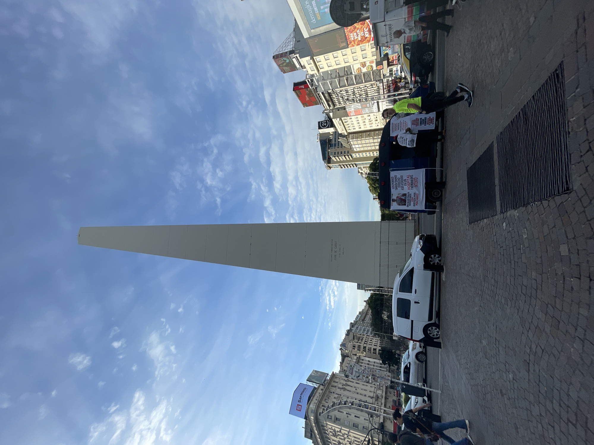 The image shows the Obelisco of Buenos Aires, Argentina, under a blue sky with light clouds. Buildings line the street in the background. In the foreground, there is a paved area with a drain. A white van is parked along the side of the road, and a smaller vehicle is towing a sign. Two individuals stand beside the sign. The sign reads "LOS POBRES TAMBIEN TENEMOS DERECHO A SER ATENDIDOS EN UN HOSPITAL". There are people walking on the sidewalk. Billboards adorn the buildings in the background.