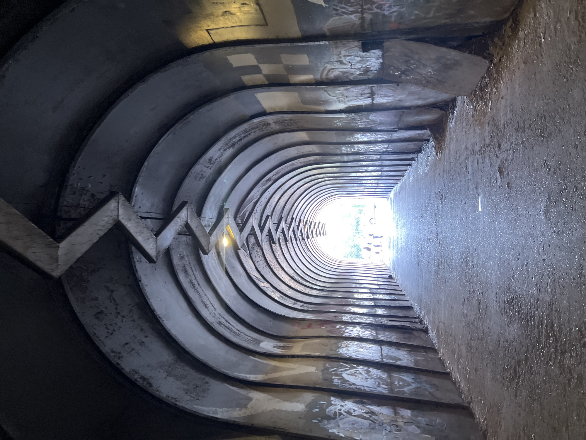 The image shows the Papagagh Tunnel, a pedestrian underpass located in Yerevan, Armenia. The tunnel is characterized by its unique ribbed construction, featuring layers of curved concrete or metal segments that create a wave-like pattern on the ceiling. Graffiti and various markings adorn the tunnel's walls and ceilings, adding to its gritty and urban aesthetic. The tunnel appears to be slightly wet, possibly due to recent rain or humidity. Bright light is visible at the tunnel's end, indicating an exit point. No people are visible in the frame.