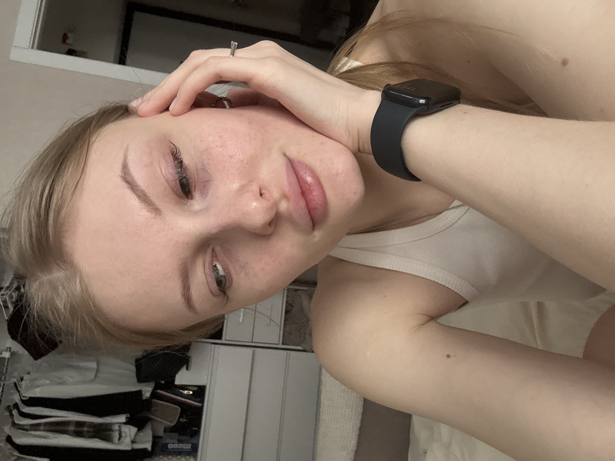 A close-up, selfie-style image shows a fair-skinned person with light brown hair. The person's right hand is raised, with fingers touching their forehead. They are wearing a ring on one finger. A black smartwatch is visible on their wrist. The person is wearing a white tank top. In the background, there is a wardrobe with clothes visible inside, a window and what appears to be bedding. The location in Perm, Russia is not evident from the visual data provided.