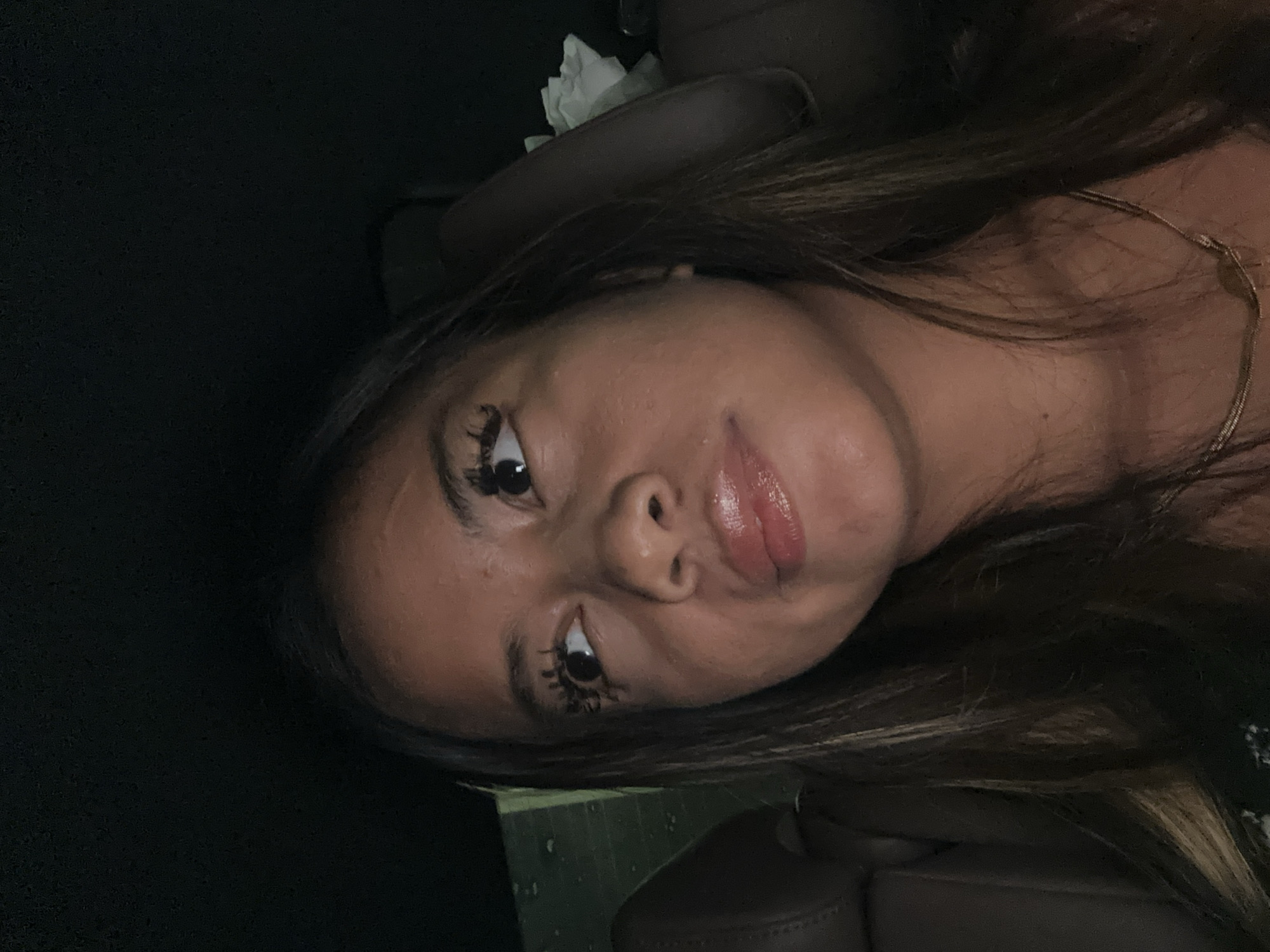 A medium close-up shot shows a woman's face at an angle. The woman has brown hair with highlighted strands, brown eyes, and full lips. She appears to be wearing makeup, including mascara on her eyelashes and lip gloss. A delicate gold necklace is visible around her neck. The background is dark, suggesting the interior of a car. The car's interior includes black leather seats and what appears to be a tissue box or crumpled tissue. The environment is dark and indistinct. The location in the city of South Jakarta, Indonesia cannot be determined from the image.