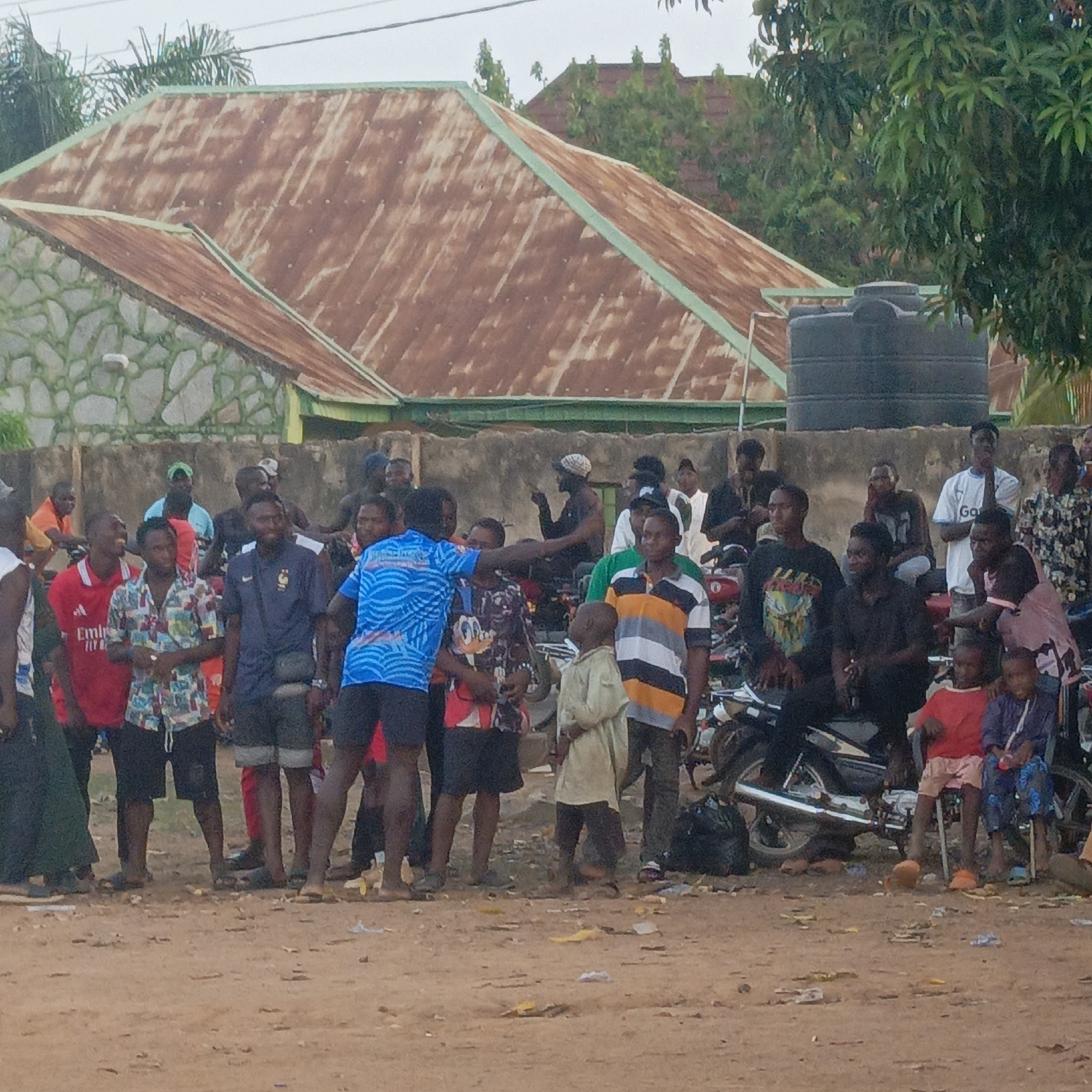 The image depicts an outdoor gathering of numerous people, primarily adult males and several children, in Akwanga, Nigeria. The scene unfolds on a dusty, unpaved ground scattered with small pieces of litter. Individuals are standing, conversing, and observing, with some seated on motorbikes parked amidst the crowd.
The subjects are dressed in casual attire, including various t-shirts and shorts. Notable clothing details include a red t-shirt with partial text "Emira TLY" and a white t-shirt displaying "Ga". A man in the center wears a blue patterned shirt with text that appears to be "MAHPEPO". Emotions conveyed are generally relaxed and observant.
In the background, residential structures are visible, characterized by rusty corrugated metal roofs, one featuring a prominent green trim, and a textured stone wall. A large, black cylindrical water tank is situated behind the wall. Lush green trees and foliage frame the upper parts of the scene. The sky is overcast, suggesting either a cloudy day or soft lighting conditions typical of early morning or late afternoon. The overall setting portrays a communal street or gathering area.