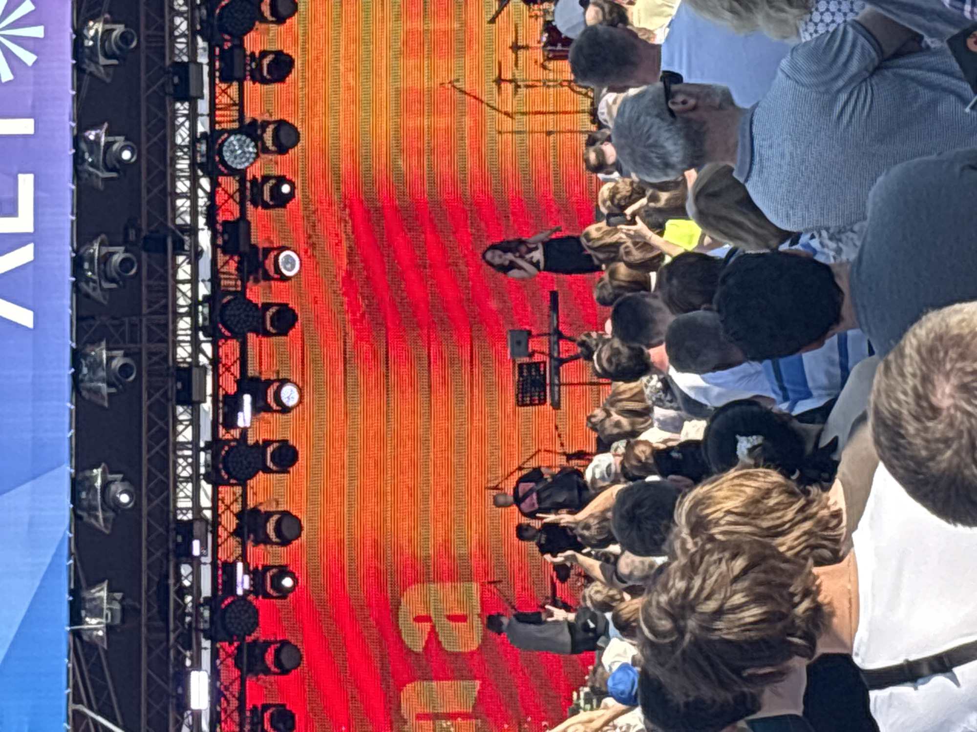 A concert is taking place on an outdoor stage. The stage has a large LED screen backdrop displaying an orange and red gradient with text that appears to read "na". Several musicians are on stage, including a female singer in the center. The audience is visible in the foreground, with many people standing and watching the performance. The lighting rig above the stage is also visible, with numerous spotlights. The overall scene suggests a lively music event.