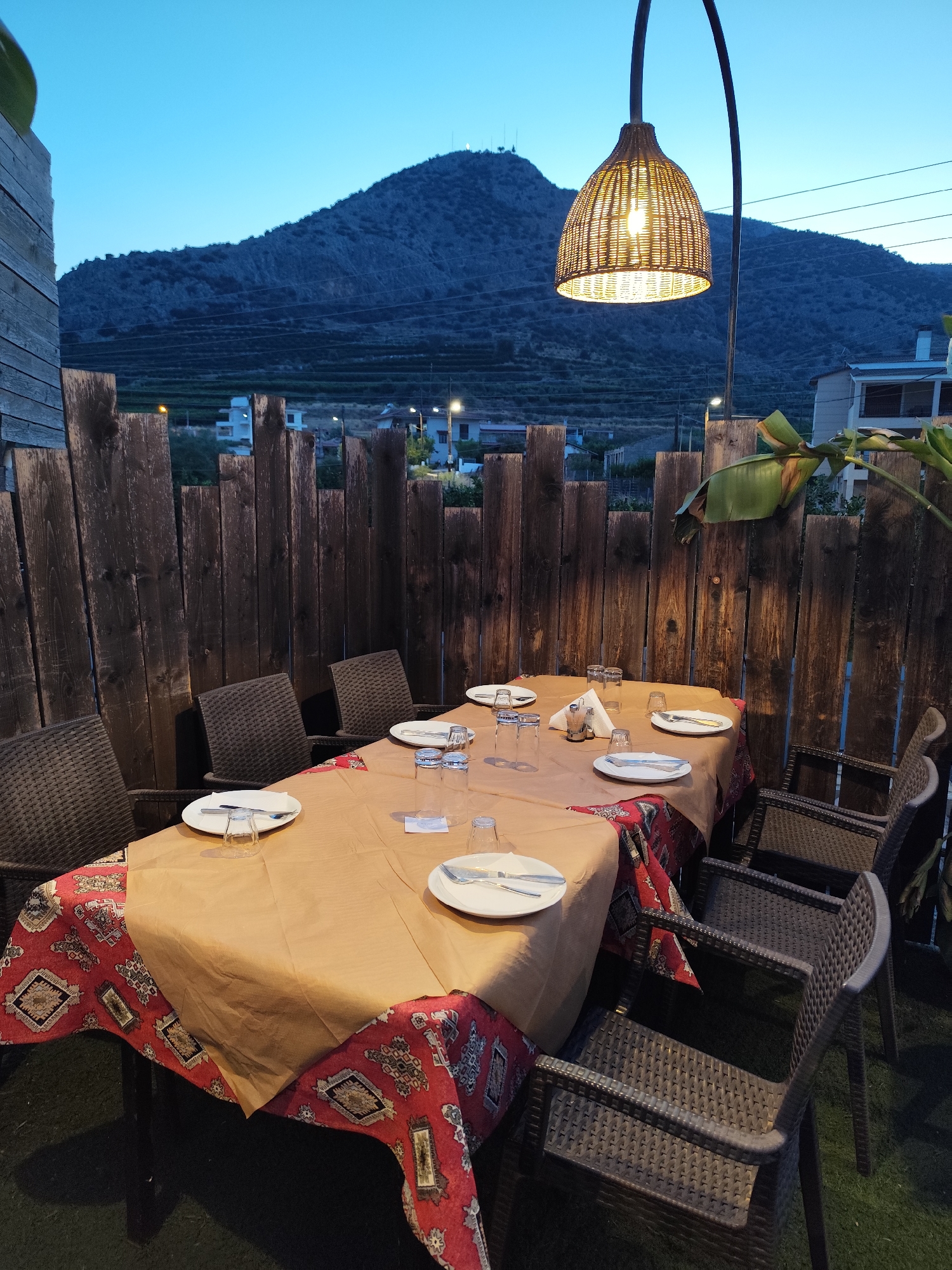 An outdoor dining setup is depicted in the Municipal Unit of Argos, Greece, during twilight. Two tables are covered with brown paper tablecloths layered over a red tablecloth with a geometric pattern. Each table is set with white plates, cutlery, glasses, and napkins. Wicker chairs surround the tables. A rustic wooden fence forms the backdrop, beyond which a hillside is visible, dotted with lights from buildings and terraces. A large, woven lampshade hangs from a curved metal pole, providing illumination. A large green plant is also present. The overall atmosphere suggests a tranquil evening setting.