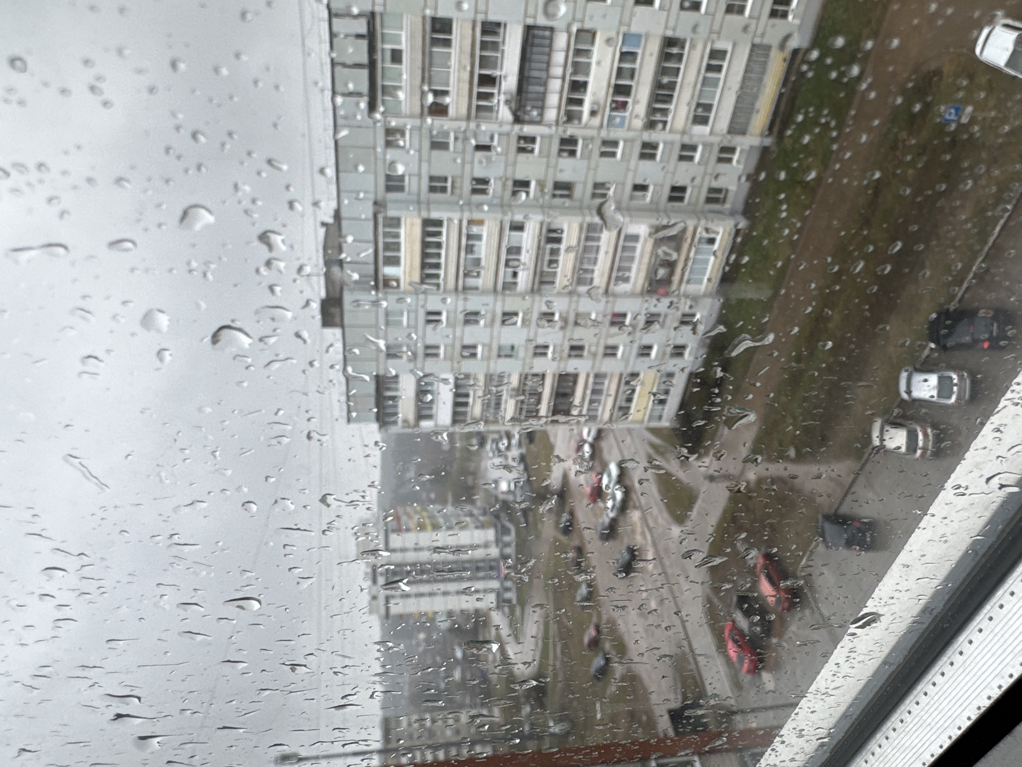 This image captures an overhead view of an urban scene in Chaikovsky, Russia, taken from behind a window heavily covered in raindrops. The foreground is dominated by myriad water droplets of various sizes, distorting the distant landscape and giving the impression of recent or ongoing rainfall.
Through the rain-streaked glass, a tall, gray residential building with numerous windows and balconies dominates the left side of the frame, typical of multi-story apartment blocks. Below, on the ground level, several vehicles are visible. These include a white car and a black car towards the right, and two distinct red vehicles parked further down the street, alongside another white car. The ground consists of paved areas and patches of dark, wet earth or grass. In the upper right section of the ground level, a small, indistinct blue rectangular sign with a white symbol is faintly visible, possibly indicating a parking or traffic regulation.
The sky, appearing in the upper left portion of the image, is uniformly overcast and light gray, confirming the cloudy and rainy weather conditions. The scene depicts a quiet, mundane moment of urban life observed from a high vantage point during a rainy day. No specific human figures or activities are clearly discernible, but the parked vehicles and residential architecture convey a sense of daily routine.