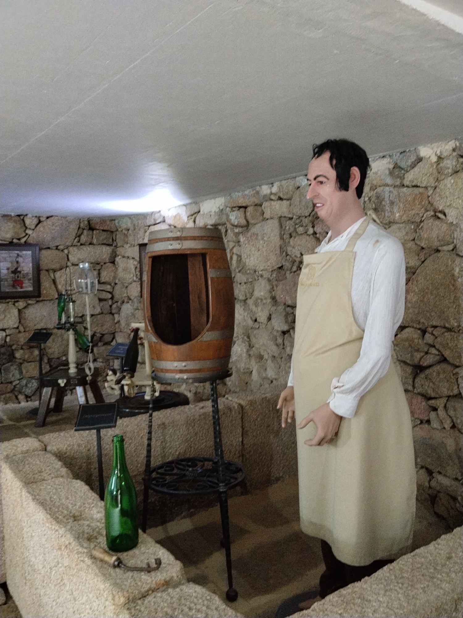 The image depicts a scene inside a stone building, possibly a museum or historical site, in Tarouca, Portugal. A life-sized mannequin stands to the right, wearing a white long-sleeved shirt and a tan apron. To the mannequin's left is a wooden barrel with a section cut out, placed on a dark metal stand. In the foreground, a low stone wall features a green wine bottle and a metal tool on its surface. The walls of the room are made of roughly hewn stones, and a framed picture hangs on the wall in the background. Behind the barrel, various other wine-related objects, including glass containers and implements, are arranged on stands. The lighting suggests an indoor setting with artificial illumination.