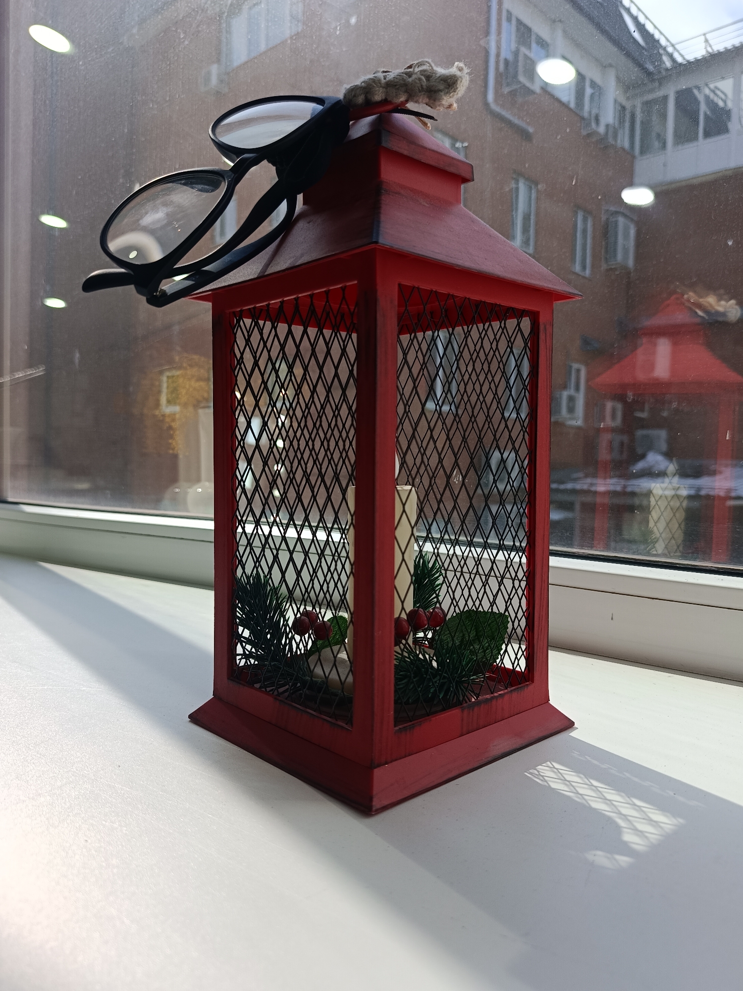A red lantern with a lattice frame stands on a white windowsill. Inside, artificial greenery, red berries, and a white candle are visible. Black glasses are perched on top of the lantern. Outside the window, a multi-story brick building is visible, along with a glassed-in walkway connecting two sections of the building. Another red lantern is visible in the reflection. The scene appears to be in Chaikovsky, Russia.