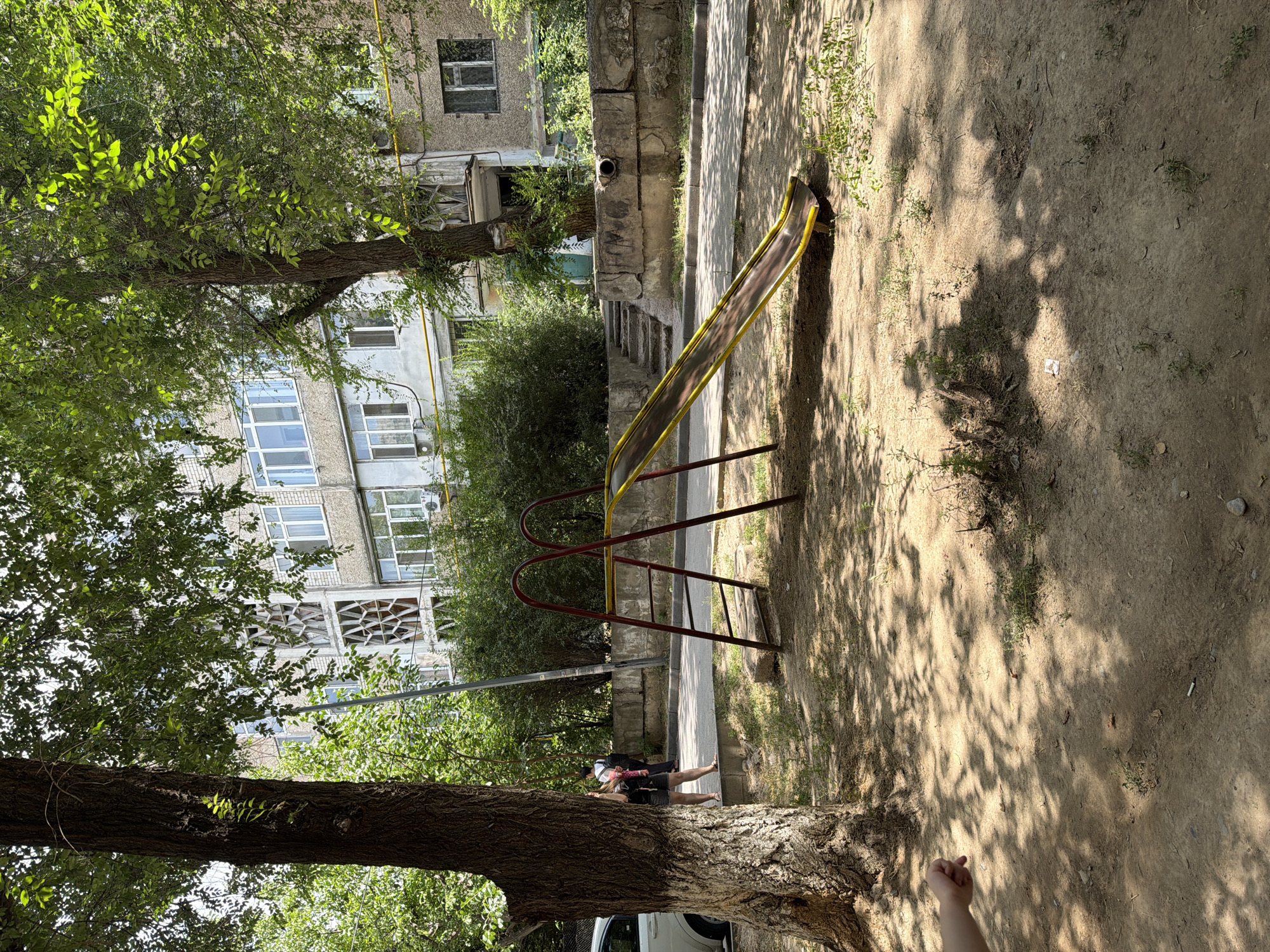 In a residential courtyard in Shymkent, Kazakhstan, a children's playground slide with red metal supports and a yellow-brown plastic slide stands on a dirt surface. Adjacent to the slide, a tree with a rough, textured bark casts dappled shadows. In the background, a multi-story apartment building with balconies and windows is visible, partially obscured by leafy green trees. A woman in dark shorts and a pink scarf is walking away from the camera on a paved path near the building. A white car is parked to the left of the frame. The scene is set on a bright, sunny day.