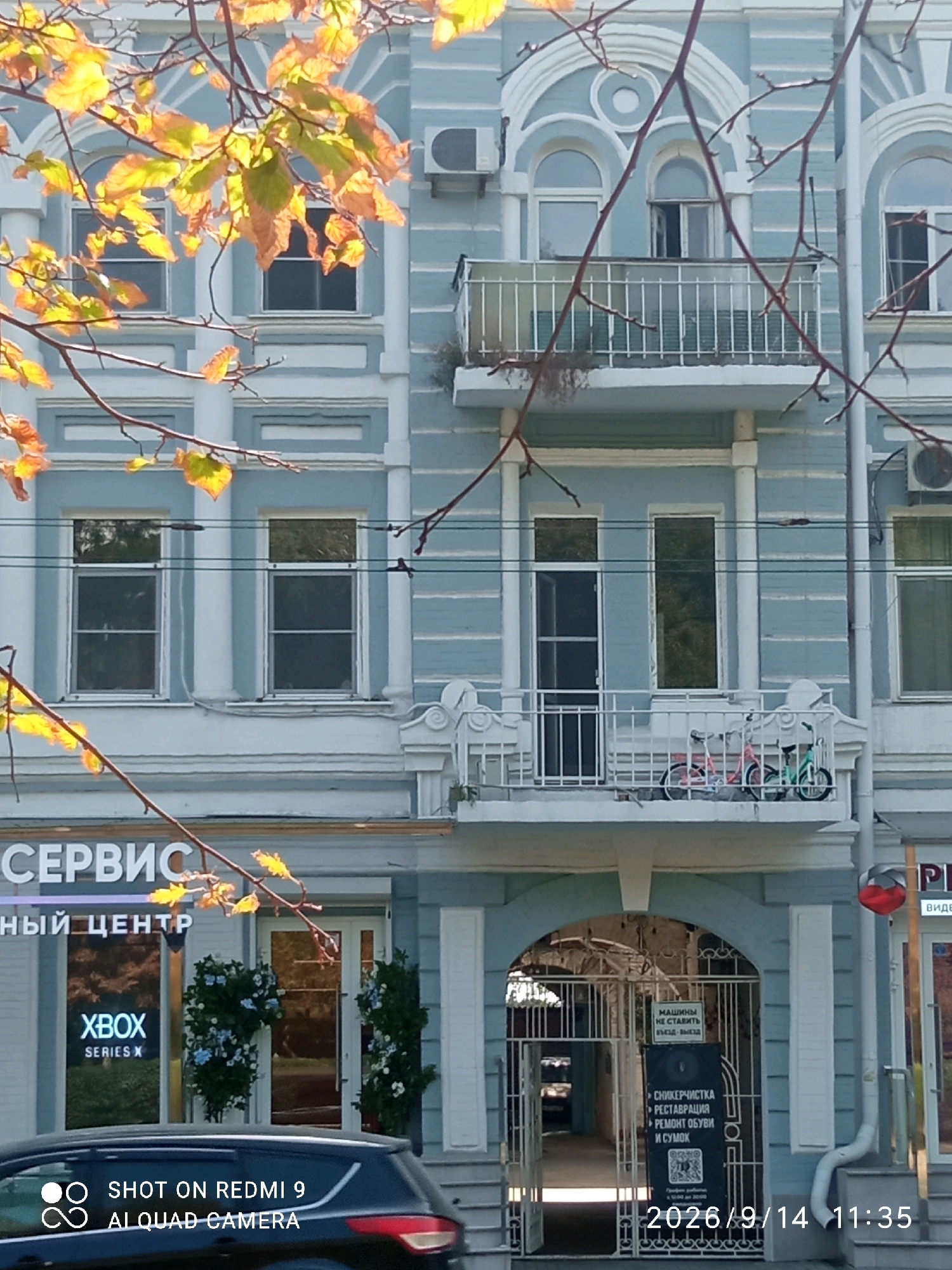 This is a full-frame, eye-level, outdoor, daylight, street-level shot of a pale blue building with white decorative molding. In the foreground, autumn leaves from a tree branch obscure the top of the building. The building features multiple stories with arched and rectangular windows, some with balconies. On the first floor, a sign reads "СЕРВИС" and below that "НЫЙ ЦЕНТР." To the left of this sign, a window displays "XBOX SERIES X." Further to the right, an arched entranceway is gated, with signs indicating services like "СНИКЕРЧИСТКА" (sneaker cleaning), "РЕСТАВРАЦИЯ" (restoration), and "РЕМОНТ ОБУВИ И СУМОК" (shoe and bag repair). A black car is parked in front of the building, with a watermark on its hood reading "SHOT ON REDMI 9 AI QUAD CAMERA." The timestamp "2026/9/14 11:35" is visible in the bottom right corner. This scene is located in Rostov-on-Don, Russia.