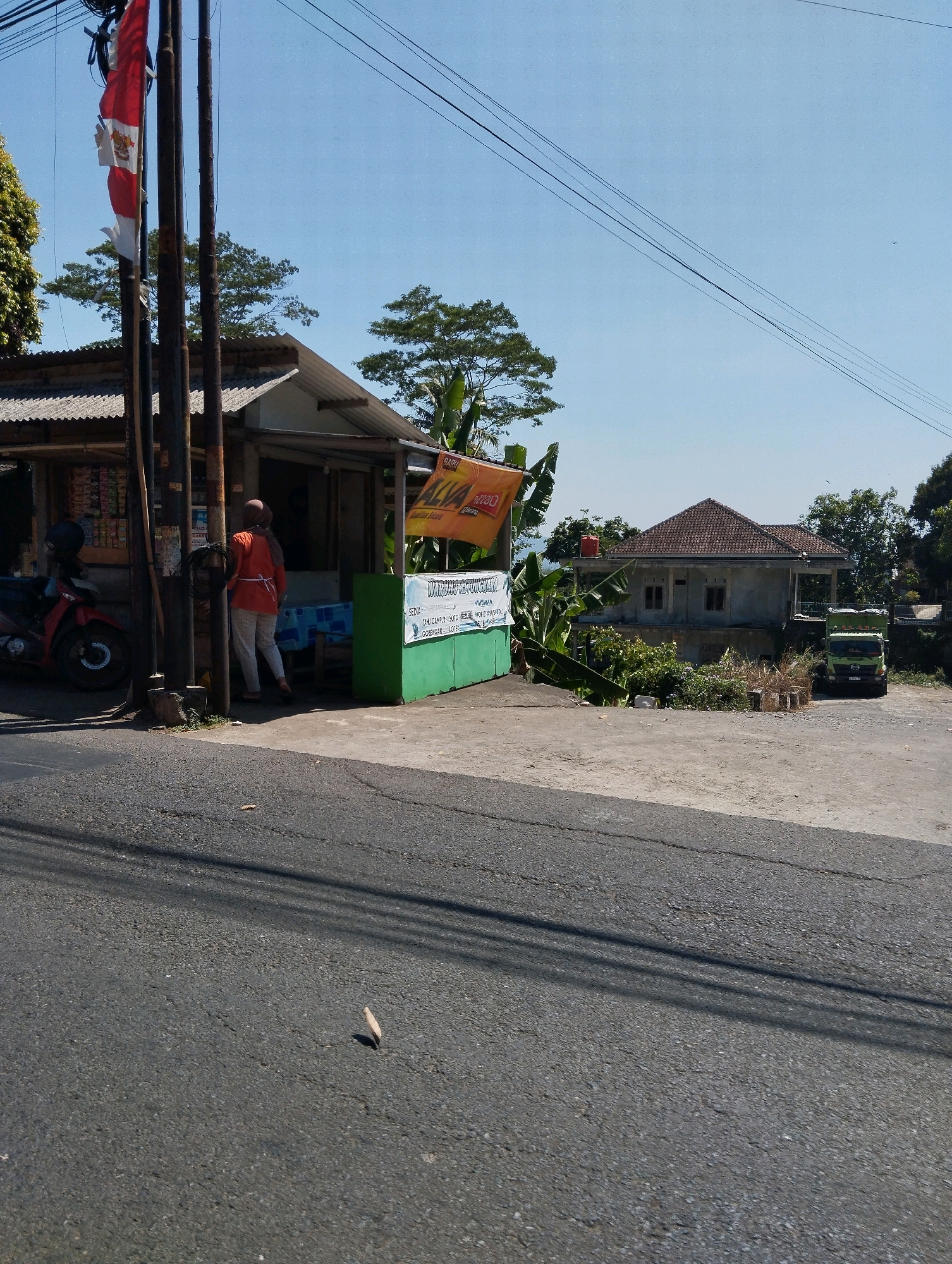 The image shows a roadside scene in Kebondowo, Indonesia, under a clear blue sky. In the foreground, a gray asphalt road occupies the lower half of the frame, with shadows of overhead power lines casting diagonal lines across it. To the left, a red motorcycle is parked next to a small building with a corrugated metal roof, which appears to be a shop or kiosk. The front of the shop displays various packaged goods. A person wearing an orange shirt and a headscarf stands near the entrance of the shop, facing away from the camera. A tall pole with a red and white Indonesian flag is also visible to the left of the shop. A green structure with signage is situated to the right of the shop, with text that appears to be in Indonesian. In the background, a light-colored house with a tiled roof is visible, flanked by trees. A green truck is parked in front of the house. The overall scene depicts a typical rural or semi-rural roadside area.