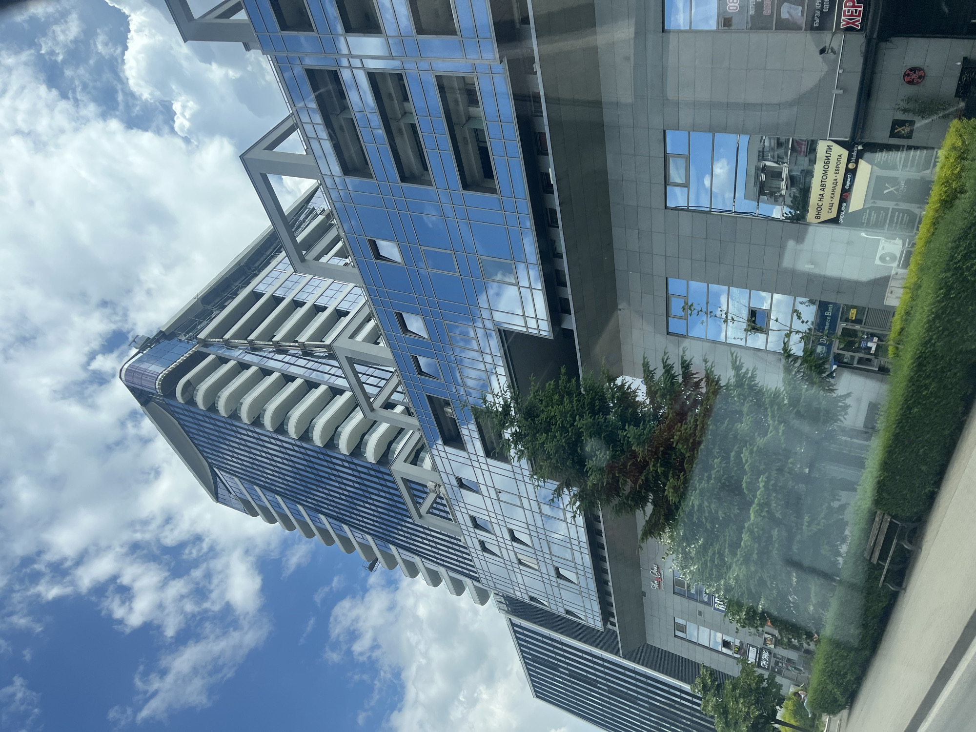 The image shows a modern, multi-story building with a glass facade and balconies. The building is tall and appears to be in an urban environment. The sky above is blue with scattered white clouds. In the foreground, there is greenery, including trees and hedges, and a wooden bench. Some text is visible on the building, including what appears to be "D Commerce Bank" and signage with text in Cyrillic. The reflection in the glass of the building shows more of the sky and possibly other buildings. The perspective is from a low angle looking up at the building.