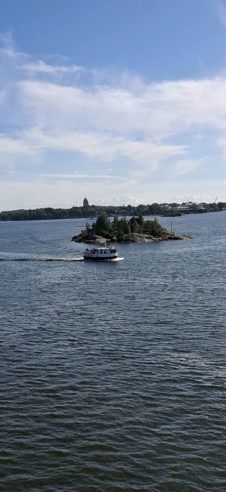 An aerial, slightly angled, full shot captures a boat on the water in Helsinki, Finland, on a sunny day. The boat is white with a black bottom and has passengers visible on deck. It is moving from left to right, creating a wake behind it. To the right of the boat is a small, rocky island covered in green trees and a few small buildings. In the background, across the water, is a long, tree-covered coastline with several buildings, including a prominent tower-like structure. The sky above is blue with scattered white clouds. The water is dark blue and choppy, with sunlight glinting off the surface. The scene appears to be a view from a ferry or a higher vantage point overlooking the sea.