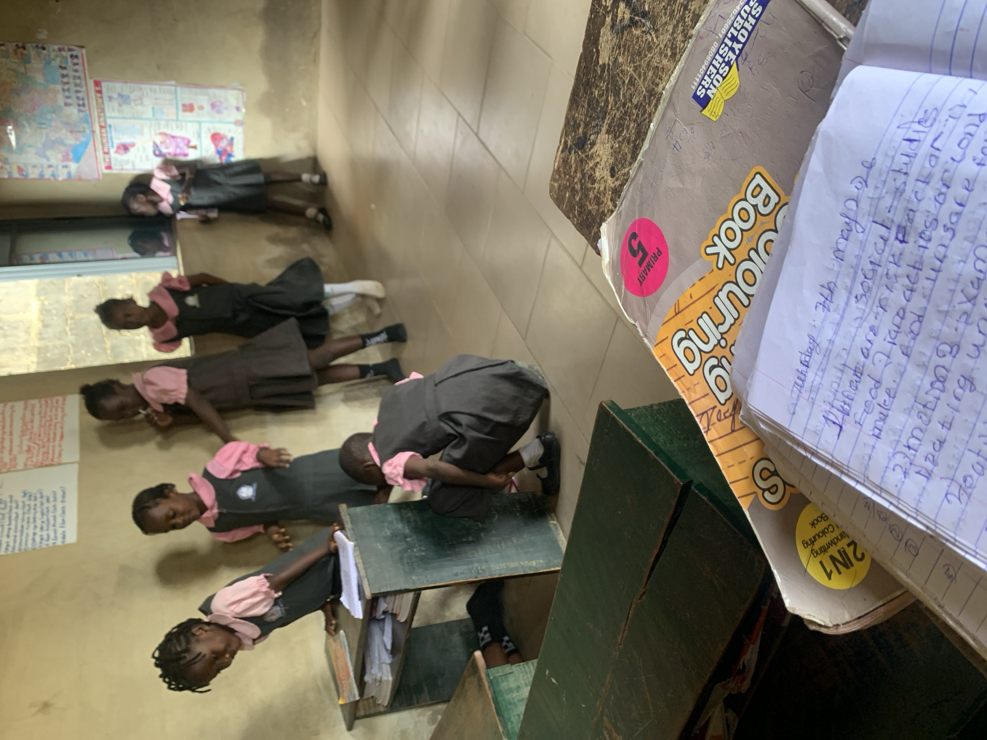 The image shows a classroom in what appears to be a school, based on the presence of desks and students. Five children, all dressed in school uniforms with pink collars and gray dresses, are lying on their stomachs on the floor. Each child appears to be looking at something on the floor, possibly reading or writing. One child is crouched on a desk.
On a table, there's a stack of books, including one with a "2IN1 Handwriting Colouring Book" label and another with a "Primary 5" sticker. The pages of a notebook are visible, filled with handwritten text. A piece of paper on the wall has writing on it, and maps hang on the wall as well.