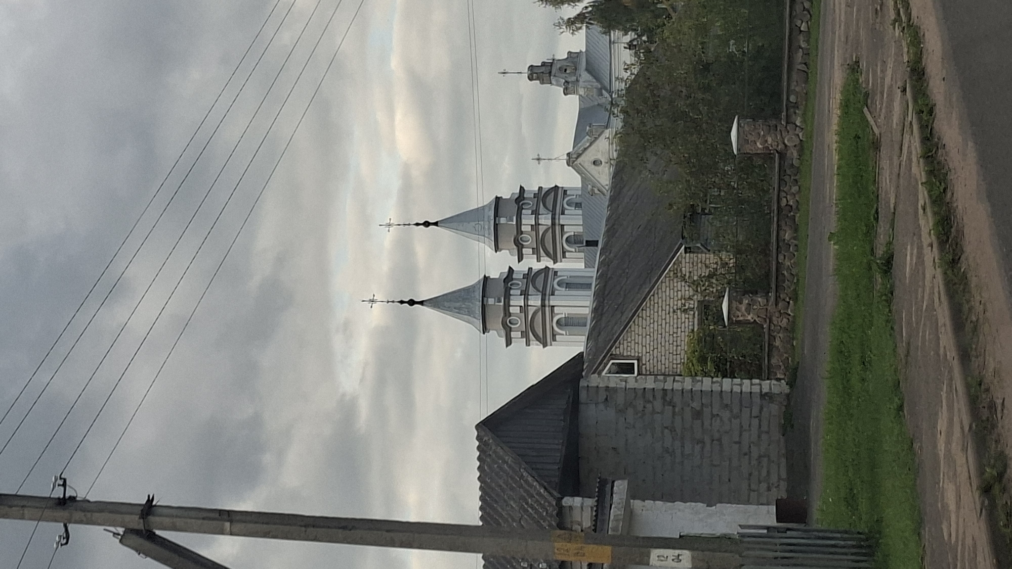 The image captures a view of a town or village in the vicinity of Сітцаўскі сельскі Савет, Belarus. In the foreground, a concrete utility pole with wires stretches across the frame. To the left, a low-rise building with a dark, sloped roof and light-colored brick walls is visible, with a segment of a gray, corrugated fence in front of it. Adjacent to this building is another structure made of brick, featuring a dark roof and a small window. A low stone wall with stone pillars forms a boundary behind these buildings. In the mid-ground and background, a large, ornate church with two prominent towers topped with crosses dominates the skyline. The sky is overcast with gray clouds. The environment appears to be rural or semi-rural, with greenery and trees interspersed with buildings. There are no people visible in the image.
