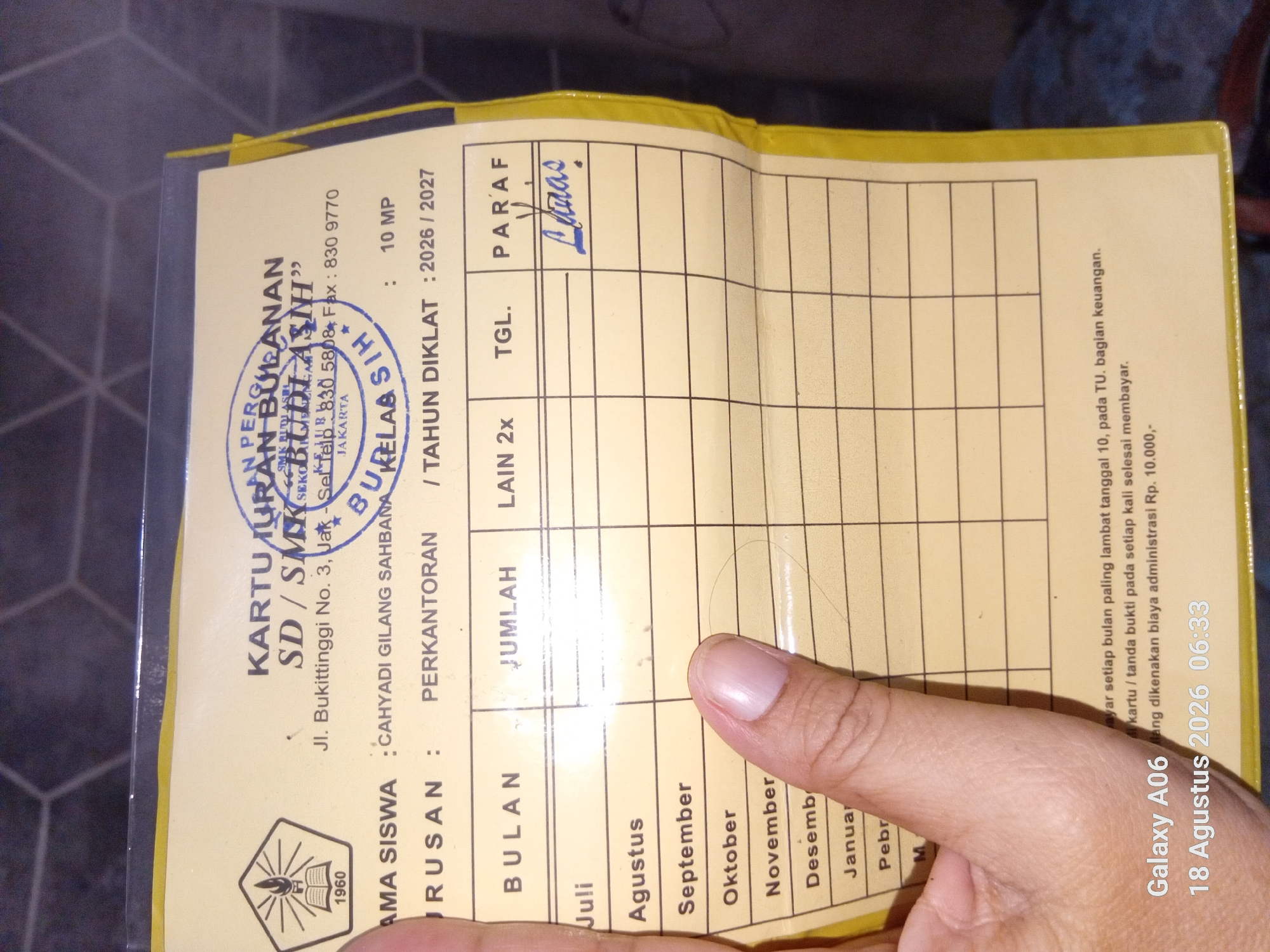 The image is a close-up, eye-level, indoor shot of a student's tuition payment card. The card is yellow with a printed grid for recording monthly payments, names of months, and columns for amount, dates, and signatures. The top of the card has official school stamps and information. The card is held by a person's hand, partially obscuring the bottom half of the card. In the background, a dark, textured surface is visible. The bottom left corner of the image has a timestamp and device name overlay: "Galaxy A06 18 Agustus 2026 06:33".
