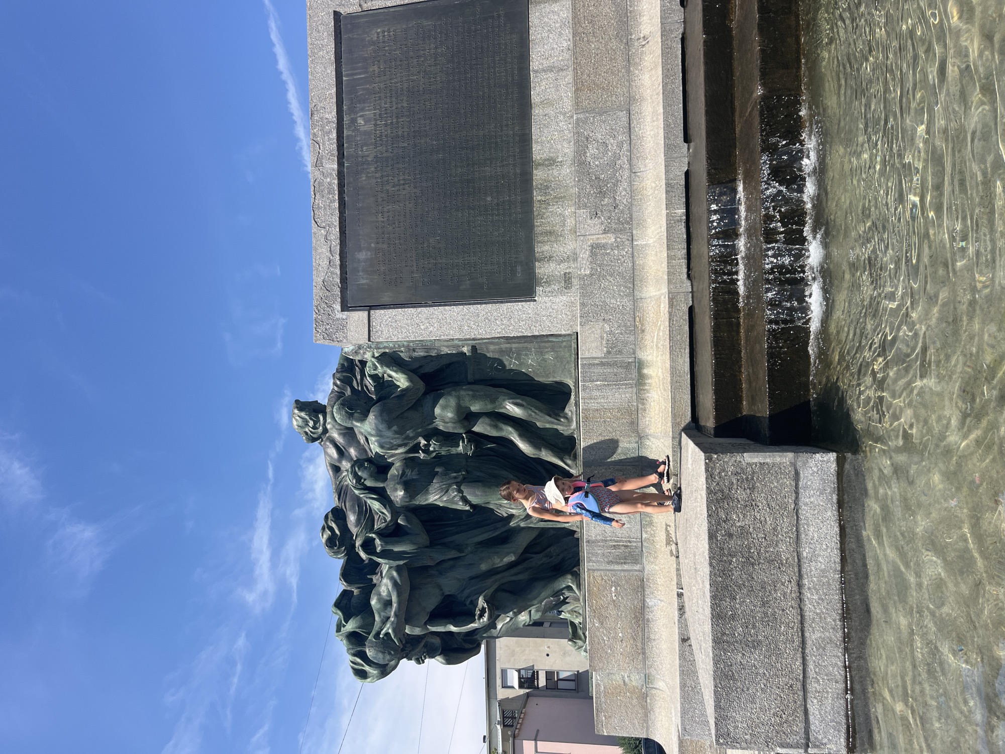 The image shows two young girls resting on the edge of a fountain in Bern, Switzerland. The fountain appears to be part of a monument with a large bronze sculpture of intertwined figures. Behind the sculpture, a granite structure has a plaque with text in French. The sky is bright blue with a few wispy clouds, suggesting a sunny day, likely in the afternoon due to the shadows. The water in the fountain is clear and calm, with sunlight glinting off its surface. The girls are dressed casually for warm weather, one wearing a hat. The overall scene is peaceful and suggests a leisurely moment outdoors. The text on the plaque is partially legible, mentioning various countries and republics.