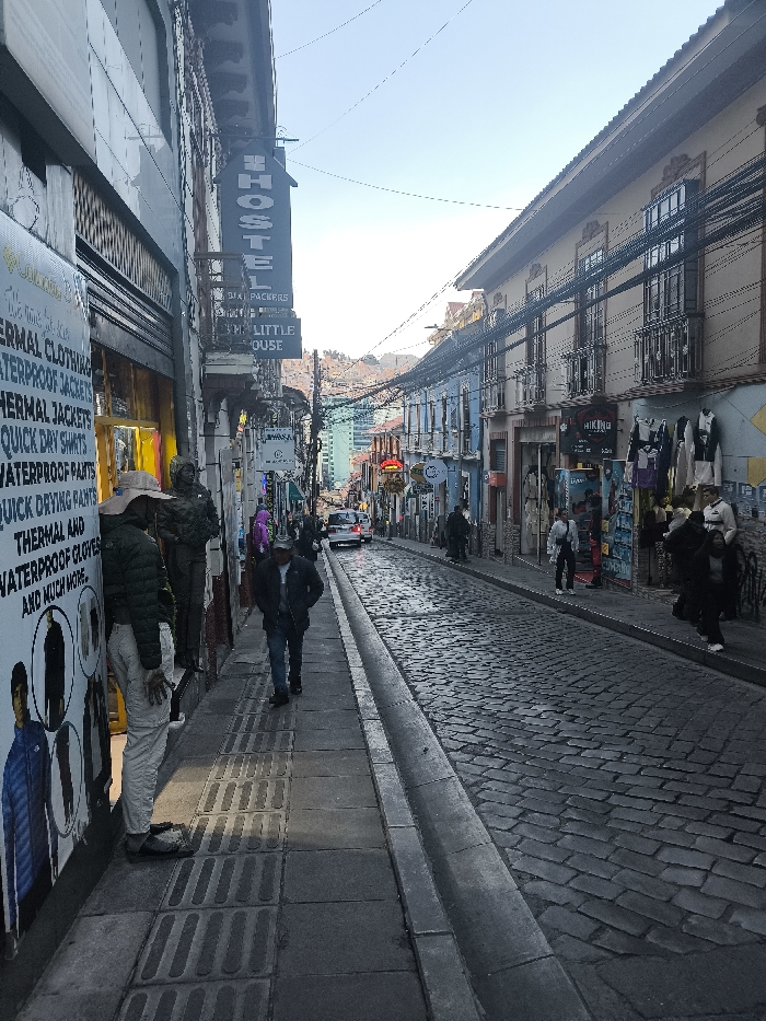 A narrow street scene features storefronts with various signs and displays. On the left, a sign advertises "thermal clothing" and "waterproof jackets." A mannequin stands nearby. Across the street, shops display clothing and travel equipment with a shop stating "Hiking." People are walking along the sidewalks and on the cobblestone street. A "Hostel" sign hangs above a building. Buildings are close together, creating a tight urban environment. Power lines stretch across the sky.