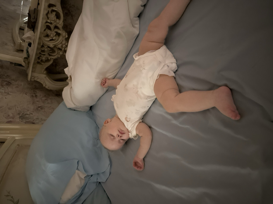 A baby lies asleep on a bed. The baby wears a light-colored onesie with spots. The bed has blue and white pillows and a gray sheet. The baby's body is positioned diagonally across the bed, with one leg extended and the other bent. Part of an ornate, white dresser is visible on the left side of the frame.