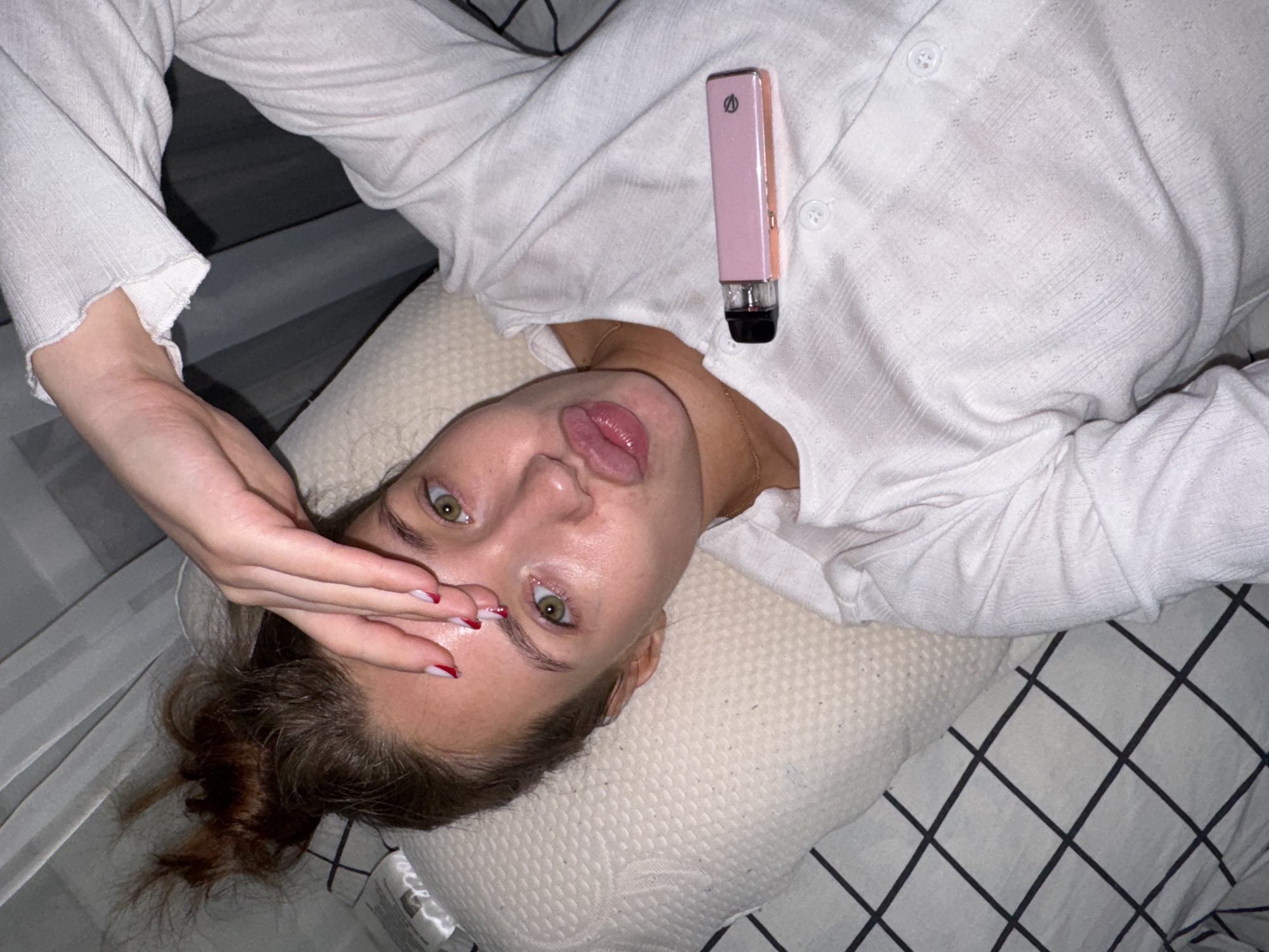 A young woman with her hair in a messy bun lies on her side on a pillow with a checkered sheet. She is wearing a white top and has her hand over her eyes. A pink vape pen rests on her chest. Her lips are pursed.