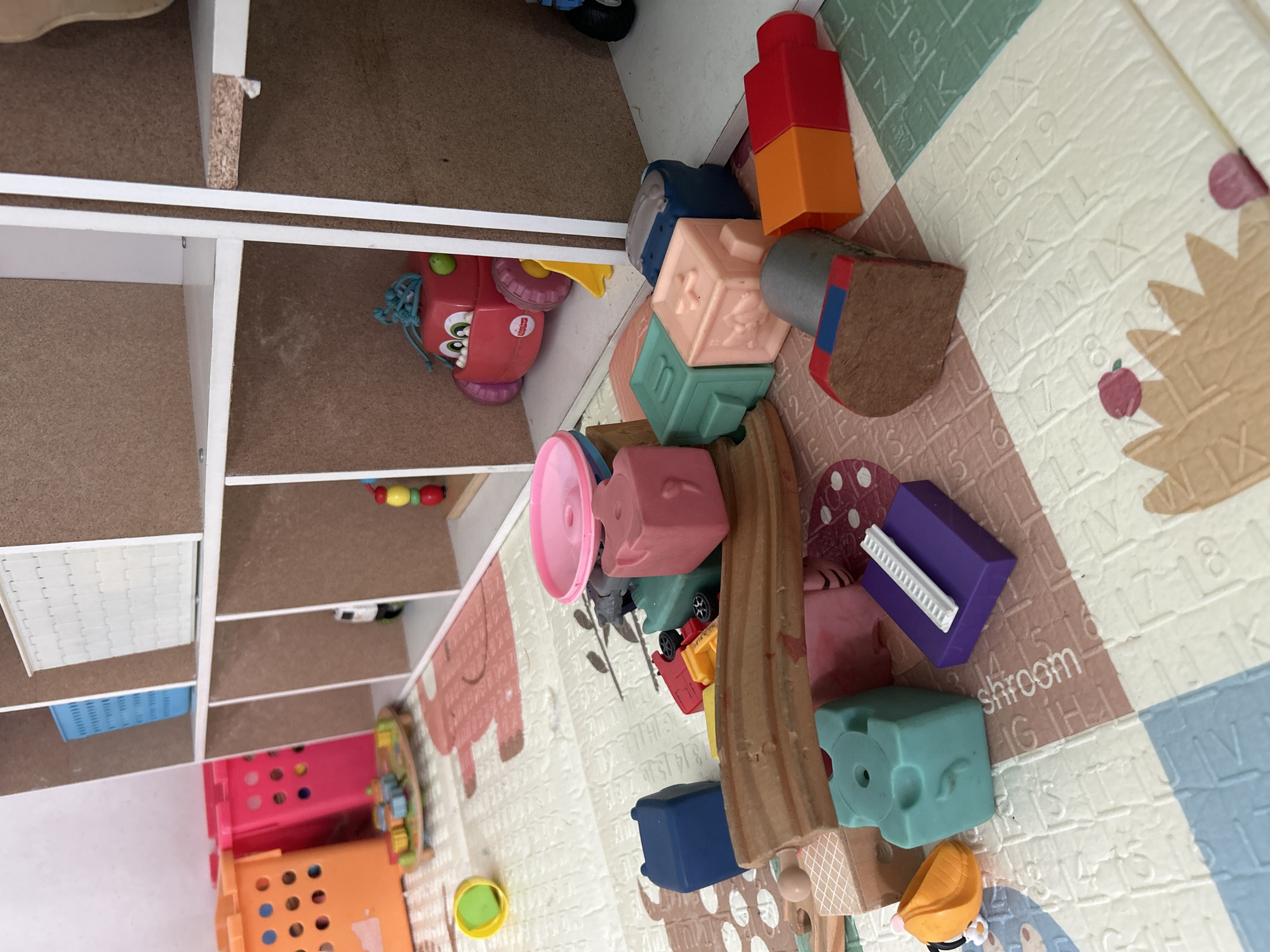 In Kuta, Indonesia, a collection of colorful children's toys is scattered on a play mat. The scene depicts a partially assembled toy train track made of wooden pieces. Several plastic blocks in various shapes and colors, including blue, green, pink, and purple, are scattered around the track. Other toys visible include a red toy car with large wheels, a yellow plastic cup, and a toy with red and orange blocks. In the background, shelves are organized with blue and white storage baskets and a smaller toy. The play mat features a pattern of letters and numbers. The lighting suggests an indoor setting, possibly a child's playroom.
