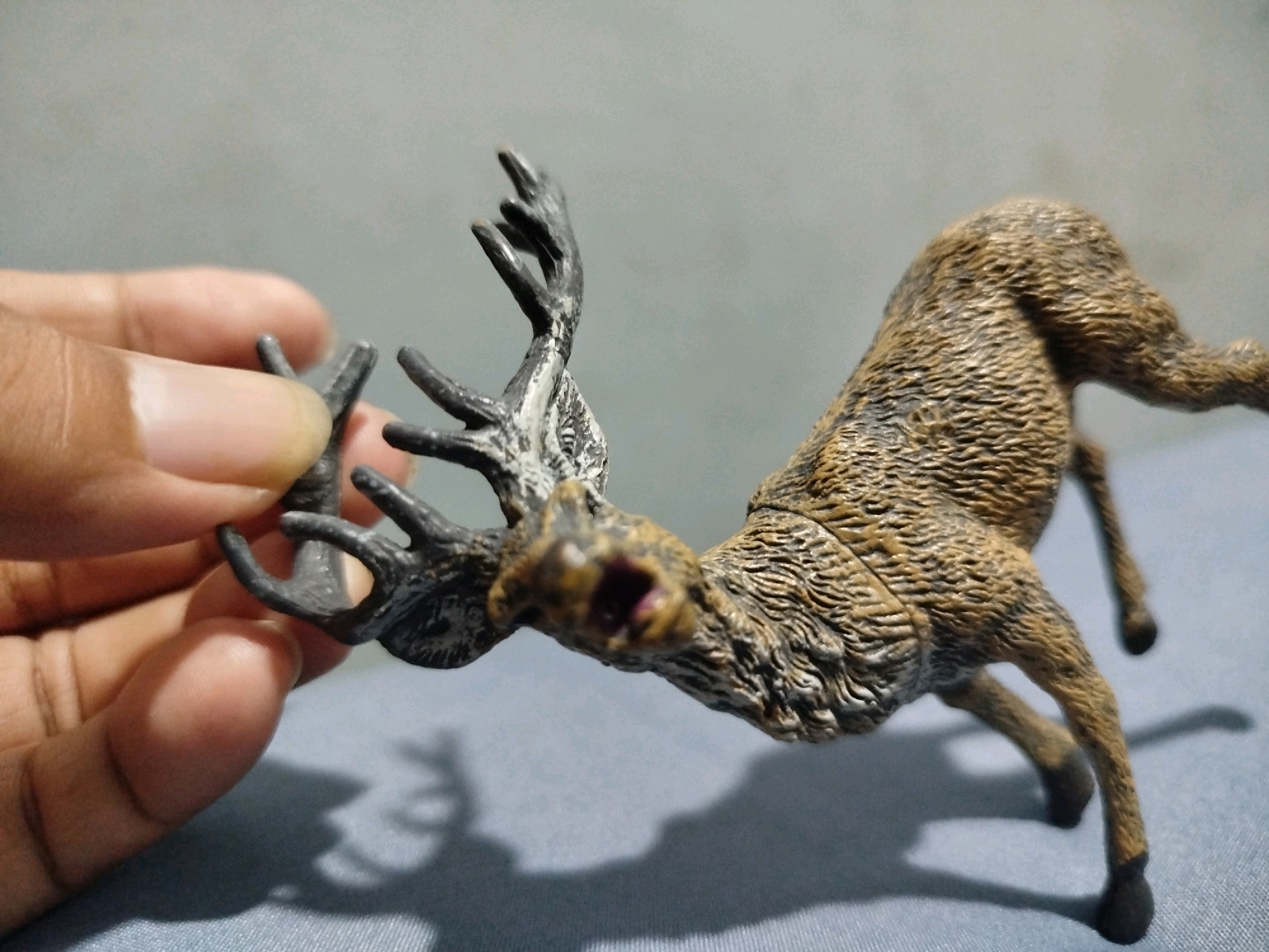 A hand is holding the antlers of a small, brown, toy deer that is lying on its side. The toy deer is on a textured surface, possibly a tablecloth. The background is a plain gray wall. The image is taken in Mojoagung, Indonesia.