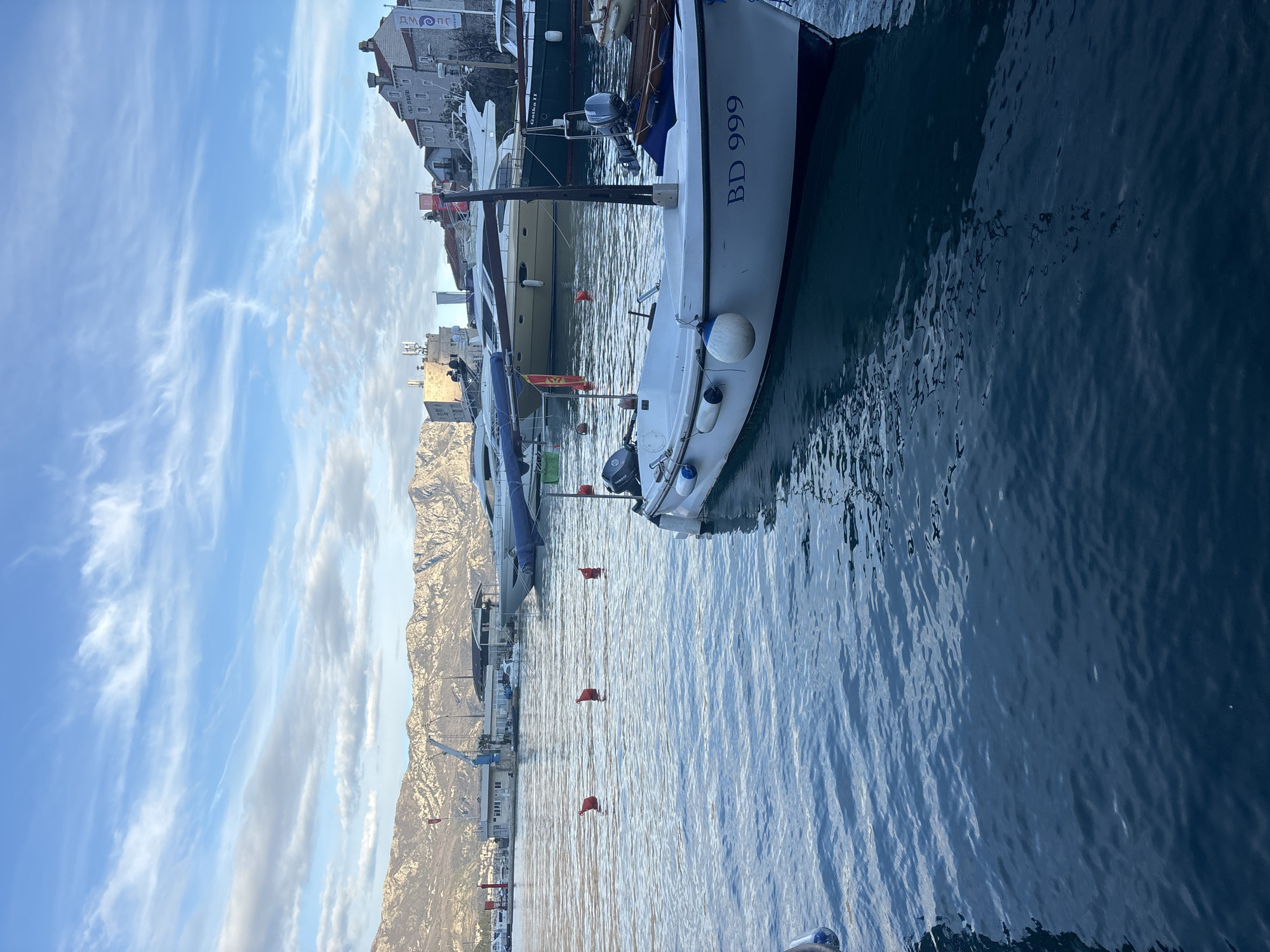 The image depicts a harbor scene in Budva, Montenegro. In the foreground is a white sailboat with the registration number "BD 999" on its hull, moored in calm, blue water. The harbor is lined with various boats and yachts, with red buoys marking the waterway. Buildings and structures are visible along the waterfront, including a building with the letters "ДМ СПС" on its roof. In the background, there is a mountain range under a partly cloudy blue sky.
