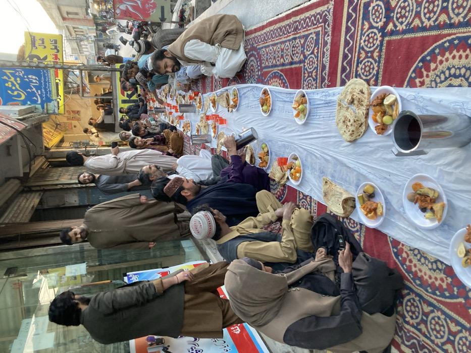 The image depicts an outdoor communal meal unfolding on a sidewalk adjacent to a commercial street in Quetta City Tehsil, Pakistan, identified by storefront signage displaying "Quetta" and shop names like "Stationery" and "Royal Hotel Kabul Biryani." Approximately 20-25 individuals are visible, predominantly adult males dressed in traditional South Asian attire, with one individual wearing a head covering. A large, patterned red, blue, and cream rug covers the sidewalk, over which a long white plastic sheet is laid as a communal dining surface.
Numerous white disposable plates are arranged along the plastic sheet, each containing a portion of mixed food items, including fried fritters and various fruit pieces. Flatbreads are also distributed along the sheet. Several silver metallic thermoses and red plastic cups are interspersed among the plates. The majority of individuals are seated cross-legged on the rug, engaged in eating and conversation. One man in the mid-ground is actively pouring liquid from a thermos into a red cup. Another individual in the immediate foreground, wearing a head covering, is holding and looking at a black mobile phone.
In the background, several men stand near the storefronts, which include both open and closed establishments with metal shutters. The street beyond shows light traffic, including motorcycles and pedestrians. The lighting suggests late afternoon or early evening.