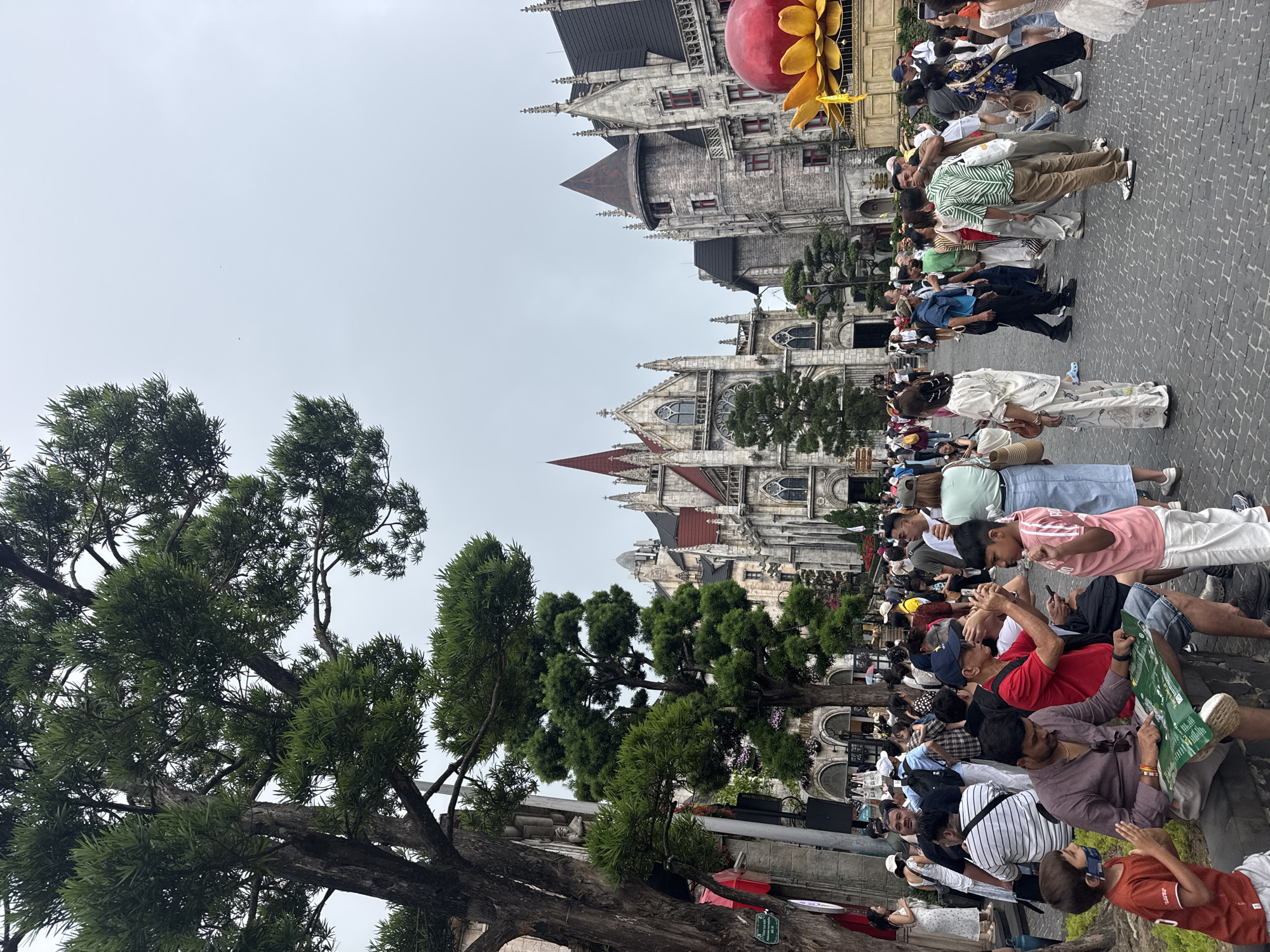 The image shows a crowded street scene in Đà Nẵng, Vietnam. Numerous people are standing or walking along a cobblestone street, with large, ornate buildings visible in the background. Trees are visible in the foreground. Many people appear to be tourists taking pictures or looking at maps. A large, artificial red apple with yellow flower is seen atop one of the buildings. The sky is overcast.