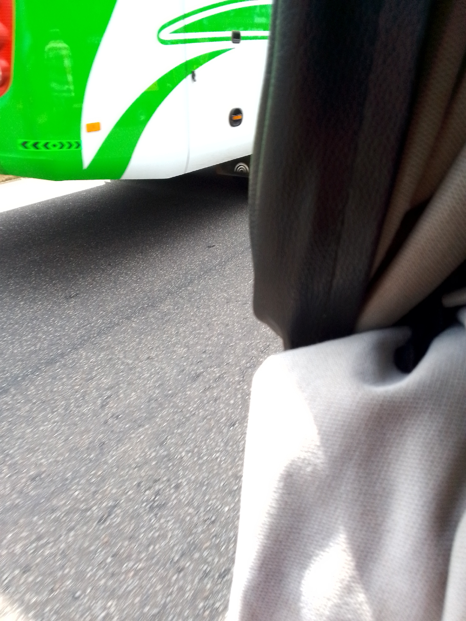 The image shows a close-up of the side of a green and white bus, as seen from inside the bus. The bus has a graphic design with curved white and green lines. There is an orange reflector and some directional arrows on the lower part of the bus. The road is visible to the left of the bus, appearing to be asphalt. A dark-colored material, likely a seat or part of the bus interior, is in the foreground on the right side of the image, along with a lighter-colored fabric, possibly a curtain. The scene is blurry, suggesting motion. There are no people or animals clearly visible.