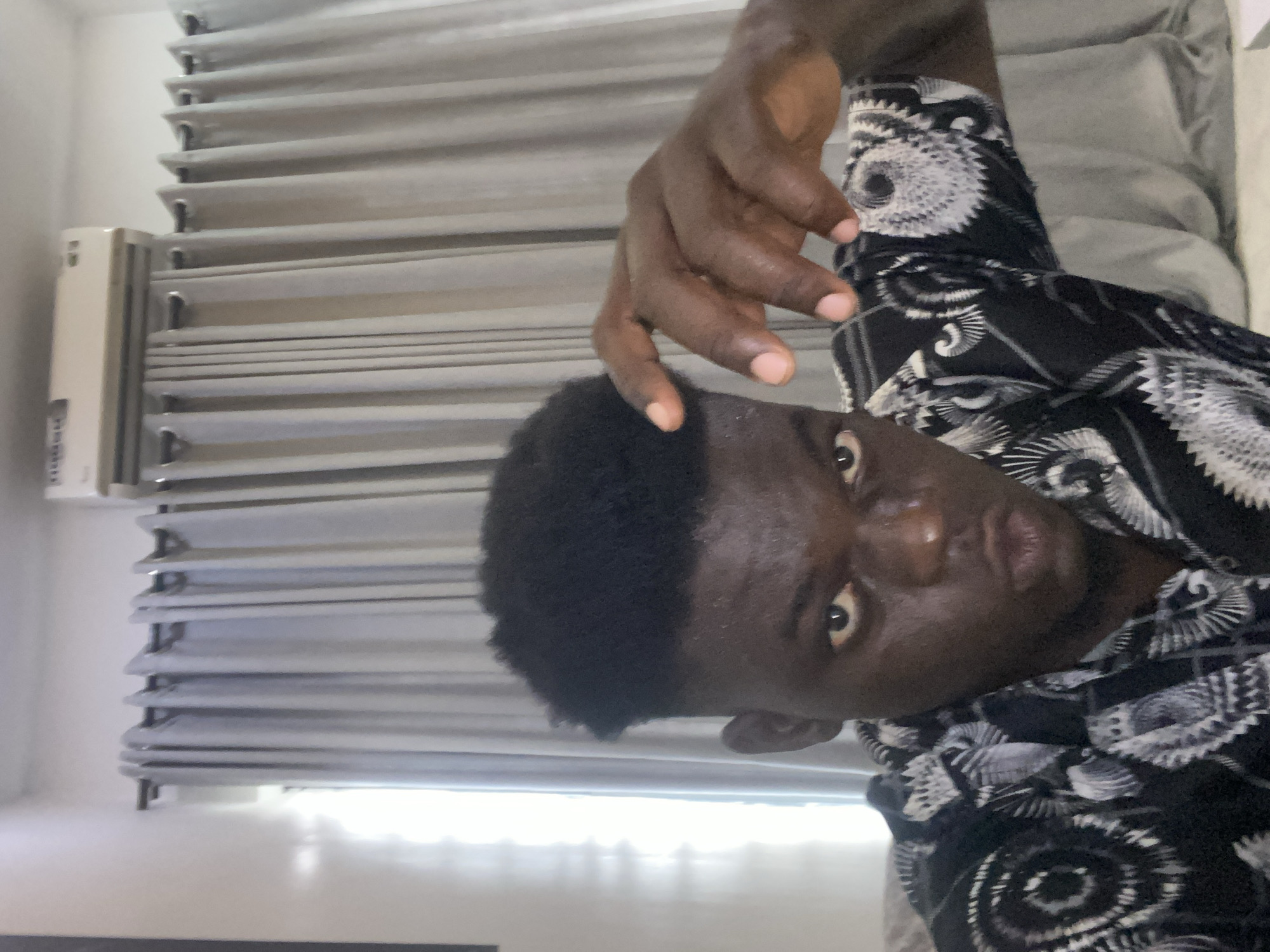 A dark-skinned person is captured in a close-up shot, appearing to be indoors with window blinds in the background. The person is wearing a black and white patterned shirt, and their hand is raised near their head. The image has been rotated, giving the impression of being taken from an unusual angle. The location is Unknown, Unknown.