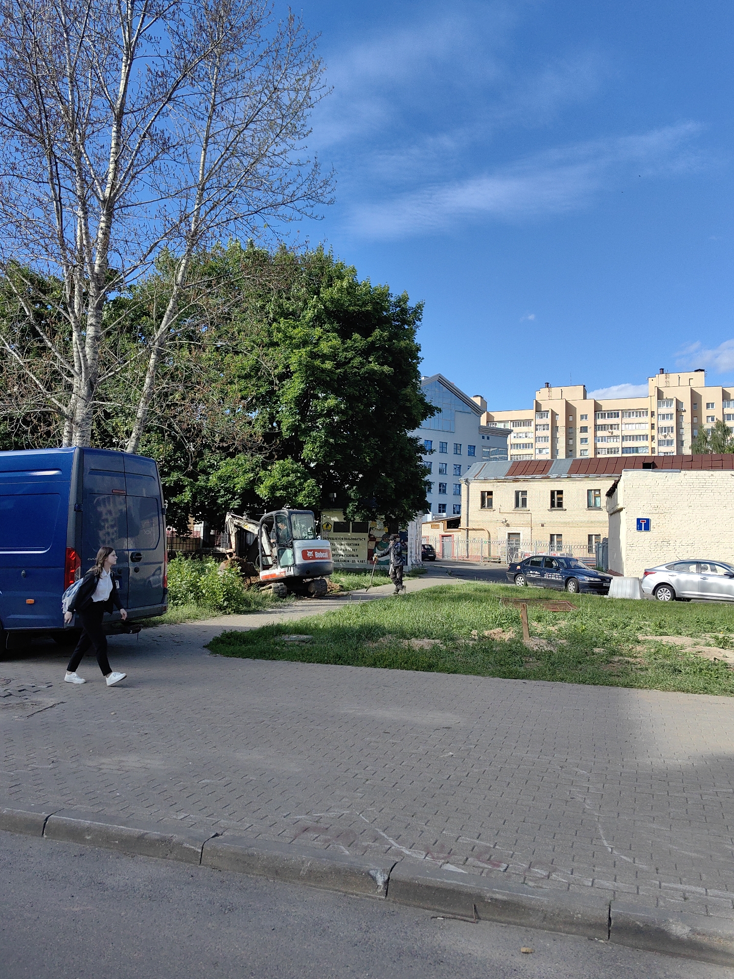 The image depicts an outdoor scene on a sunny day, likely in an urban environment in Minsk, Belarus. In the foreground, a paved sidewalk runs along a street, with a concrete curb separating it from the asphalt road. A blue van is parked on the left side of the image, and a young woman with a backpack is walking away from it on the sidewalk. Further down the sidewalk, a small excavator is positioned in a grassy area next to a dense green tree. In the background, various buildings are visible, including a light-colored multi-story residential building and a lower, yellowish brick building with a red-tiled roof. Several cars are parked on the street or in a lot near the buildings. The sky is clear blue with a few wispy clouds. The scene suggests ongoing construction or maintenance work in the area.