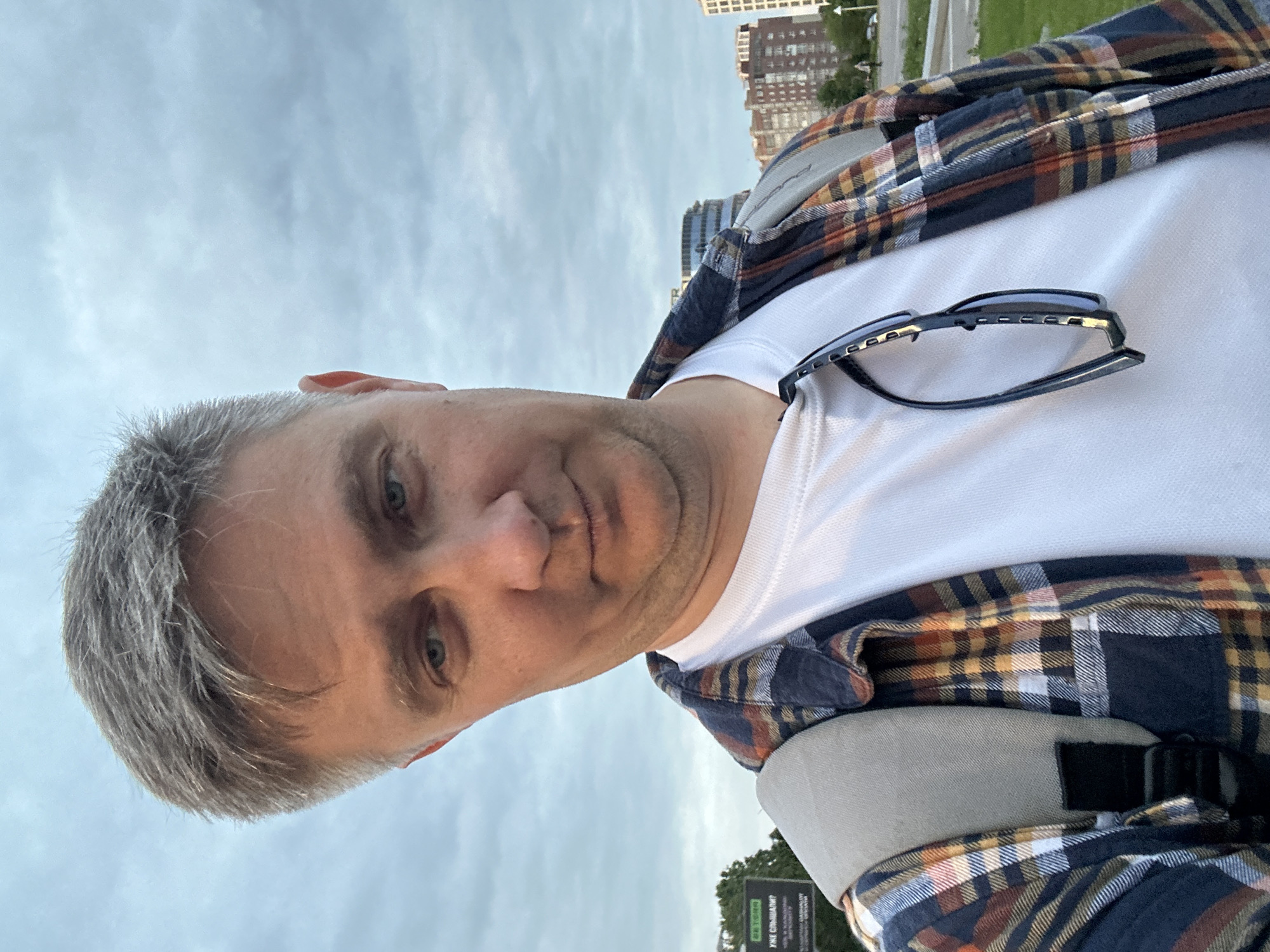 A male individual with short, greyish-brown hair and blue eyes is depicted in a close-up, chest-up shot, looking directly at the viewer, suggesting a selfie. He wears a white crew-neck t-shirt layered under an open long-sleeved plaid shirt featuring a pattern of dark blue, orange, brown, and white. Sunglasses with dark frames hang from his t-shirt collar. A light-colored backpack strap is visible over his left shoulder.
The background reveals an urban environment under an overcast, light blue-grey sky. To the viewer's left, a large green billboard is partially visible, displaying white Cyrillic text, including "24 ЧАСА" (24 HOURS), "БЕТОН" (CONCRETE), and "УЖЕ СЛЫШАЛИ?" (HAVE YOU HEARD YET?). Behind the billboard are trees. To the viewer's right, a modern city skyline features several buildings, including one with a distinctive curved, multi-story glass facade, and a multi-story reddish-brown residential building. Green grass occupies the immediate background, leading to a visible paved road or pathway in the distance. The architecture and Cyrillic text on the billboard are consistent with the city of Minsk, Belarus.