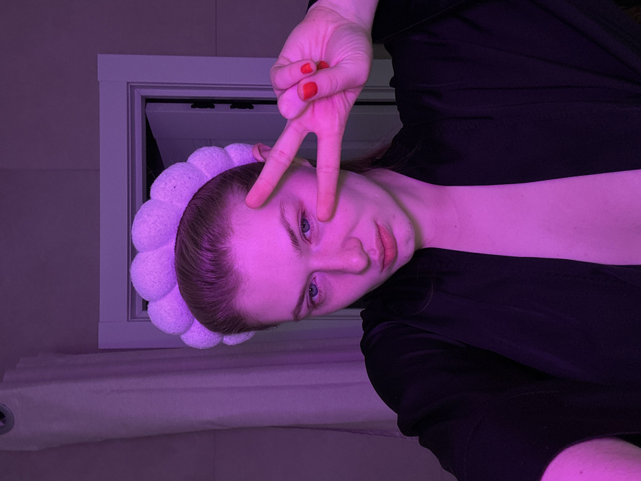 A young woman poses with her head tilted, bathed in purple light. She wears a fluffy white headband and a black top. Her hand is raised, forming a peace sign over her left eye. Her nails are painted red. Behind her is a white window frame. The overall aesthetic is a blend of casual and playful.
