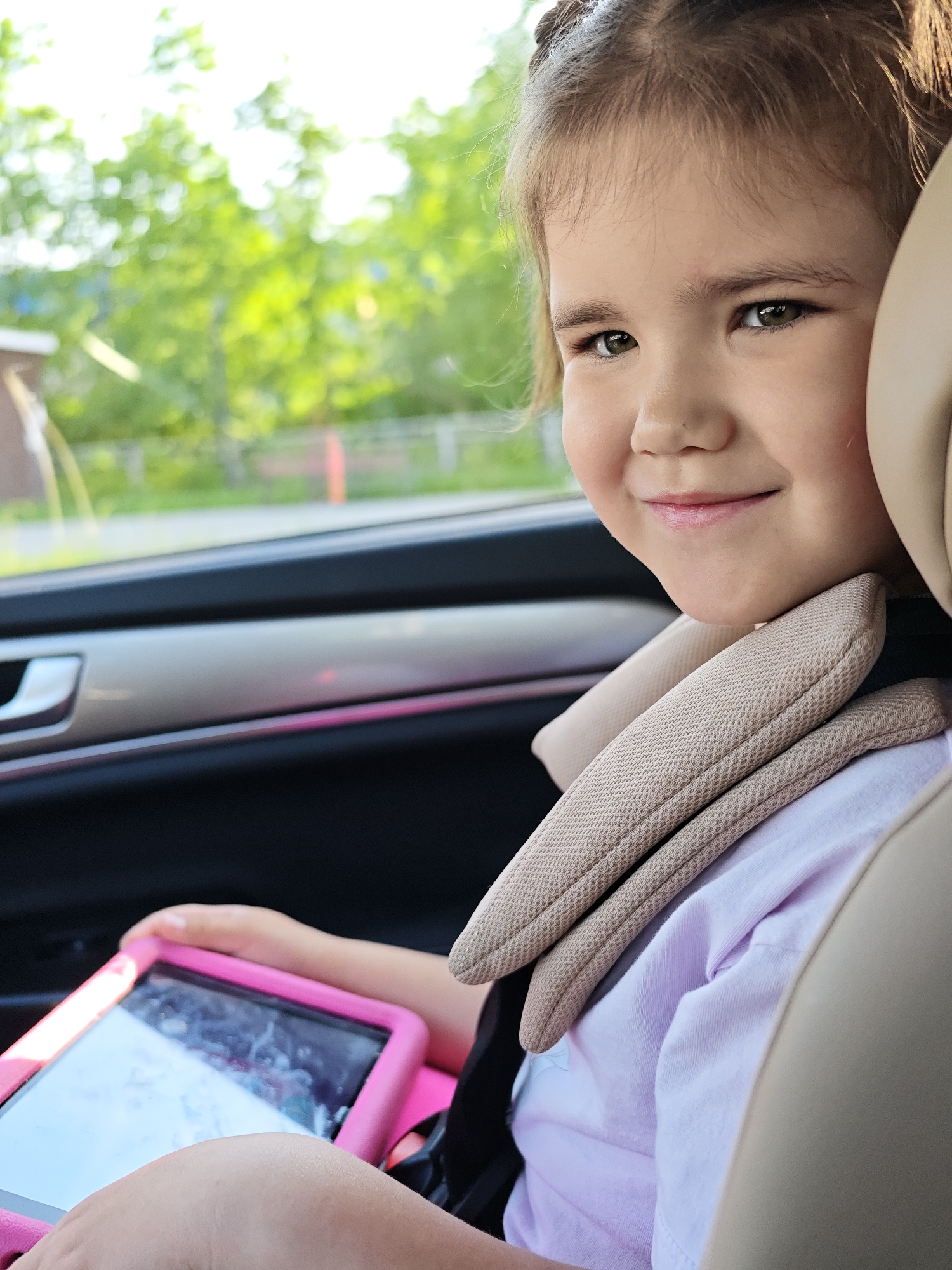 A young girl is seated in a car, wearing a safety harness. She's holding a tablet with a pink case. Outside the car window, there's foliage. The picture was taken in Chaikovsky, Russia.