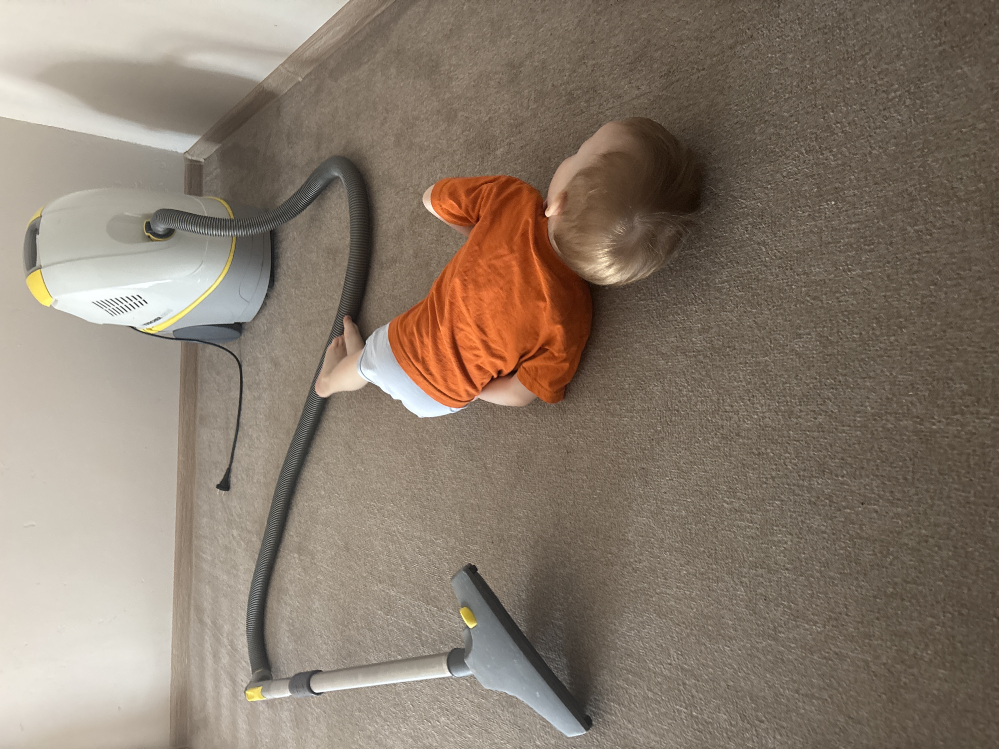 A young child with blonde hair, wearing an orange shirt and light-colored bottoms, is lying on a carpeted floor. Next to the child is a vacuum cleaner, with the hose extending toward the child. The vacuum cleaner is primarily light gray and yellow. The power cord for the vacuum cleaner is visible. The scene is indoors, and a wall is visible in the background. There is no explicit evidence in the image to confirm the location as Ферма, Russia.