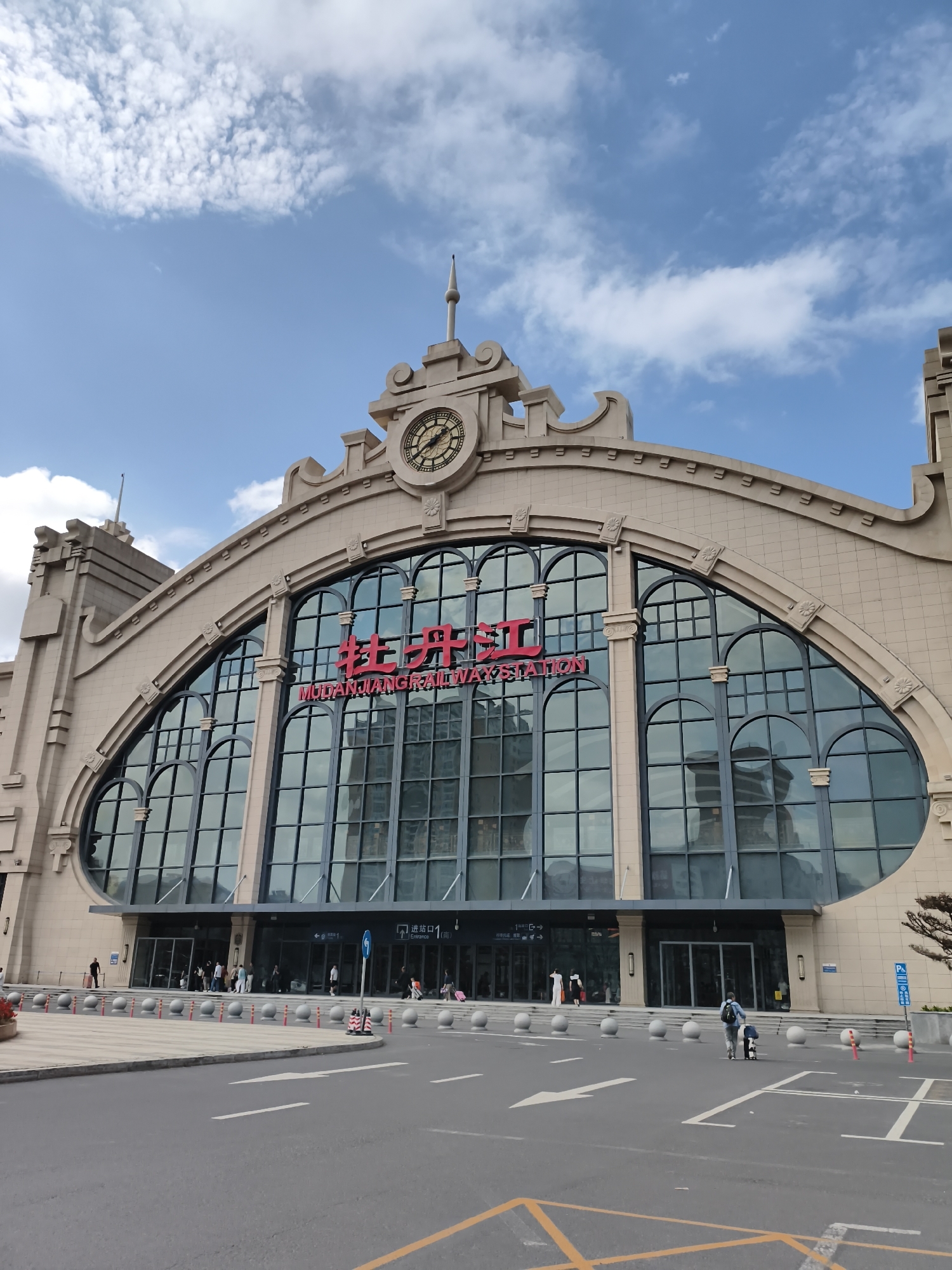 The image shows the exterior of Mudanjiang Railway Station in Mudanjiang, China. The station's facade features a large, arched glass window with red lettering that reads "牡丹江" (Mudanjiang) and "MUDANJIANG RAILWAY STATION". A clock is prominently displayed above the arched window, and the building has a classical architectural style with stone cladding. The sky above is blue with scattered white clouds. In front of the station, there is a paved area with a road, white lane markings, and yellow directional arrows. Several people are visible walking on the paved area or near the entrance of the station, some with luggage. Bollards line the edge of the paved area. The overall scene depicts a functional transportation hub on a clear day.