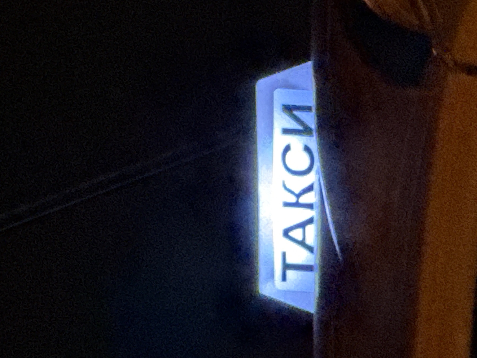 The image shows a close-up of a illuminated taxi sign that reads "ТАКСИ" in Cyrillic letters, a word that translates to "TAXI" in English. The sign is a light blue color and appears to be mounted on the roof of a vehicle. The background is dark and out of focus, suggesting the photograph was taken at night or in a dimly lit environment. There is a faint hint of the vehicle's body visible beneath the sign, appearing to be a dark, textured surface. The overall impression is that of a taxi waiting for passengers in a city. The presence of Cyrillic script suggests this location is in a country that uses this alphabet, such as Bulgaria. Varna, Bulgaria is a coastal city and a popular tourist destination, and it is plausible to find taxis with such illuminated signs there.