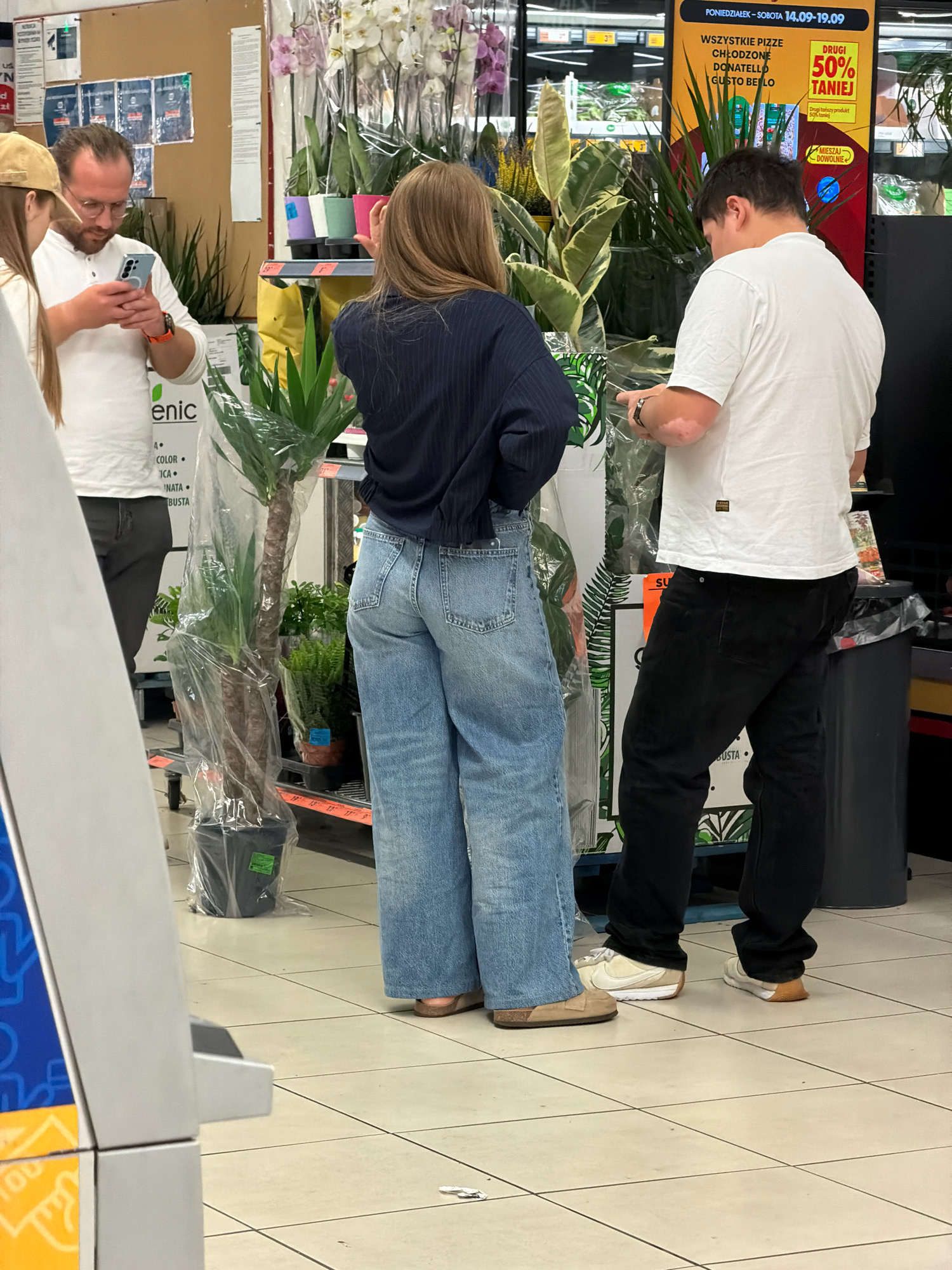 The image depicts a scene inside a retail store, likely a supermarket or a garden center, in Wrocław, Poland. In the foreground, a woman with long brown hair is standing, facing away from the camera, wearing a dark blue long-sleeved shirt and light blue wide-leg jeans. To her left, a man with glasses and a beard, wearing a white polo shirt and dark trousers, is looking at his phone. Another man, wearing a white t-shirt and black trousers, stands to the right of the woman, also looking at his phone. The environment is characterized by shelves stocked with various plants, some wrapped in plastic, and promotional signage. The floor is tiled with a grid pattern. The overall lighting suggests an indoor retail setting.
